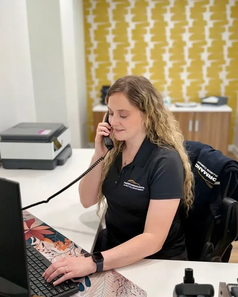 A woman in a black polo shirt sits at a white desk, speaking on a telephone while typing on a computer.