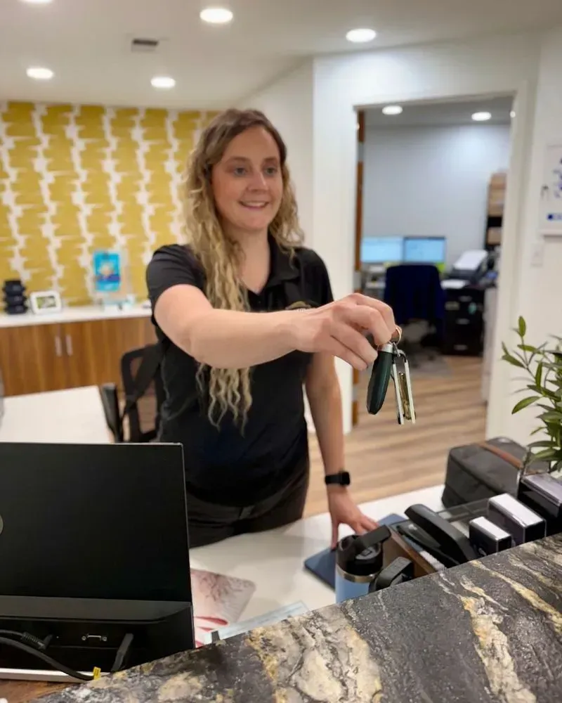 A person behind a front desk extends their hand, offering a set of car keys while smiling in a brightly lit office.
