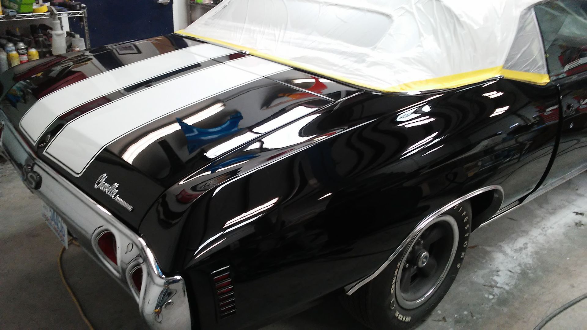 A black Chevrolet Chevelle convertible with white racing stripes on the trunk in an auto body repair shop.