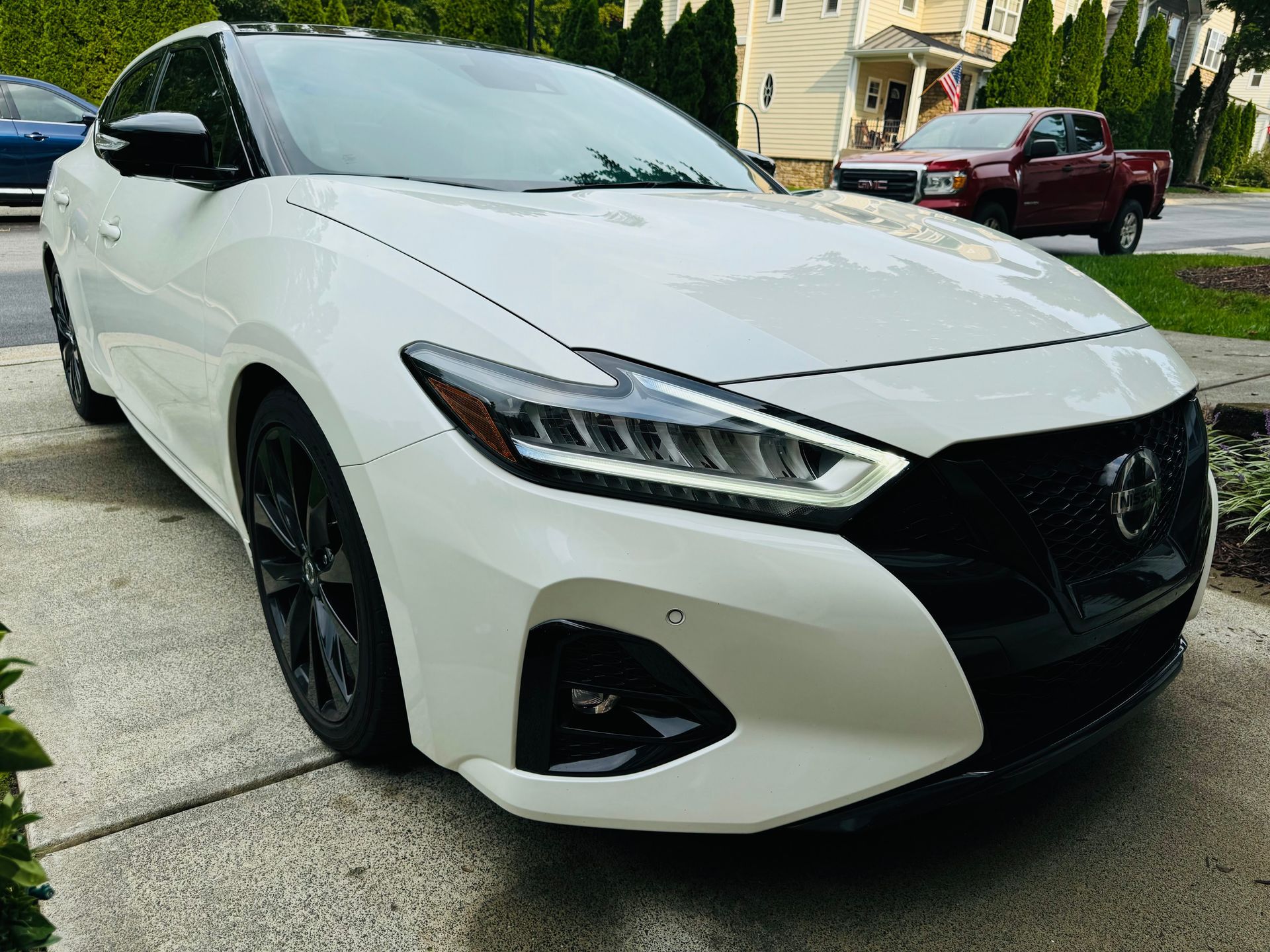 A white Nissan Maxima sedan with black rims parked on a residential driveway on a sunny day.