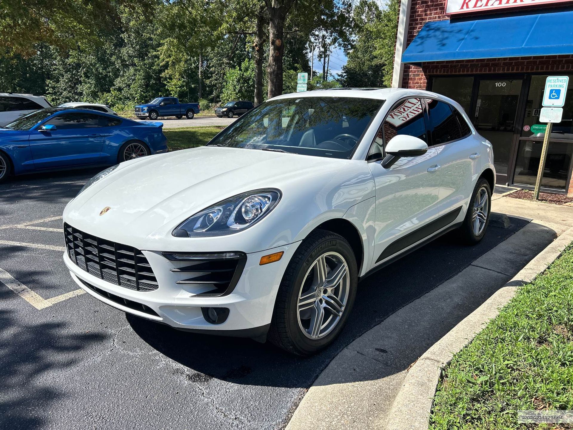 A white Porsche Macan SUV parked in a paved lot next to a building with a blue awning.