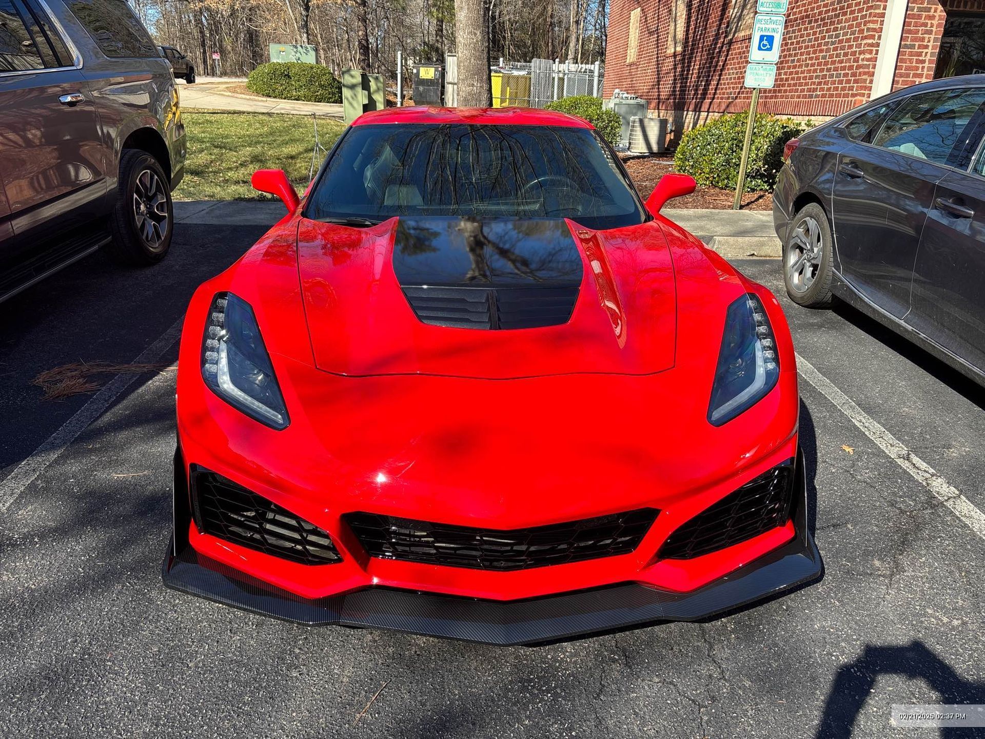 A bright red sports car with a black hood section and carbon-fiber front splitter parked in a parking lot.