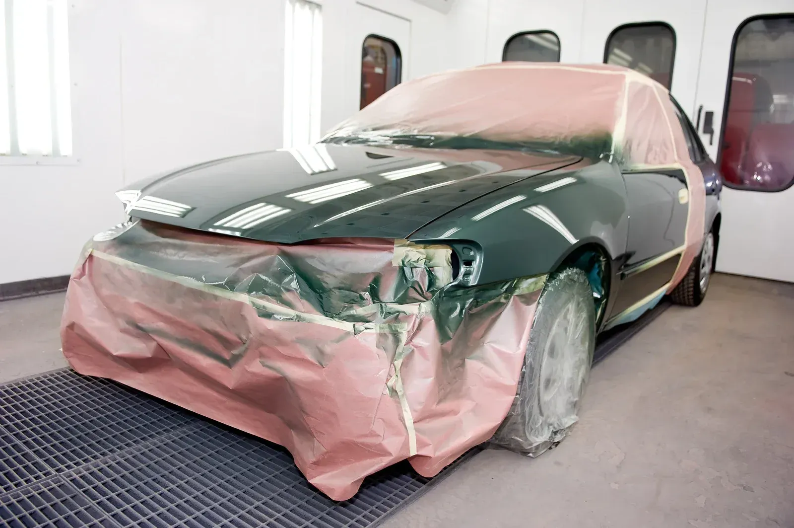 A technician in a white protective suit and respirator sprays red paint onto a car panel in an auto body shop.