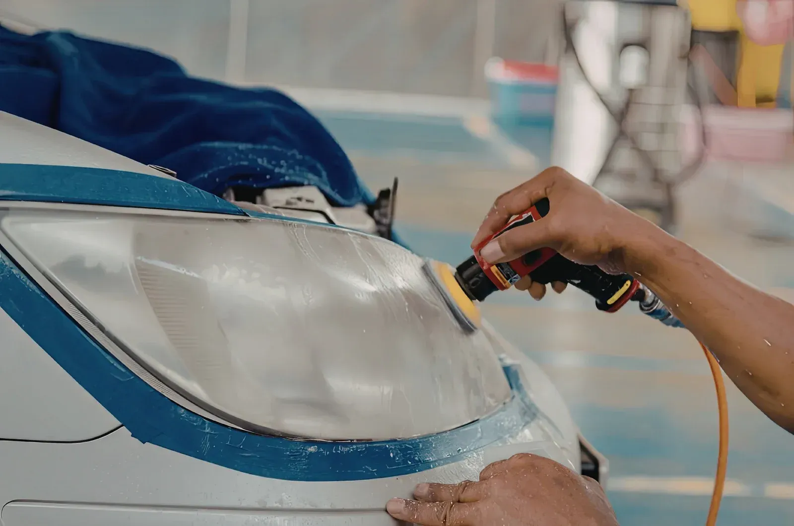 A person uses a power tool with a yellow pad to polish the surface of a car headlight, which is taped off with blue tape.