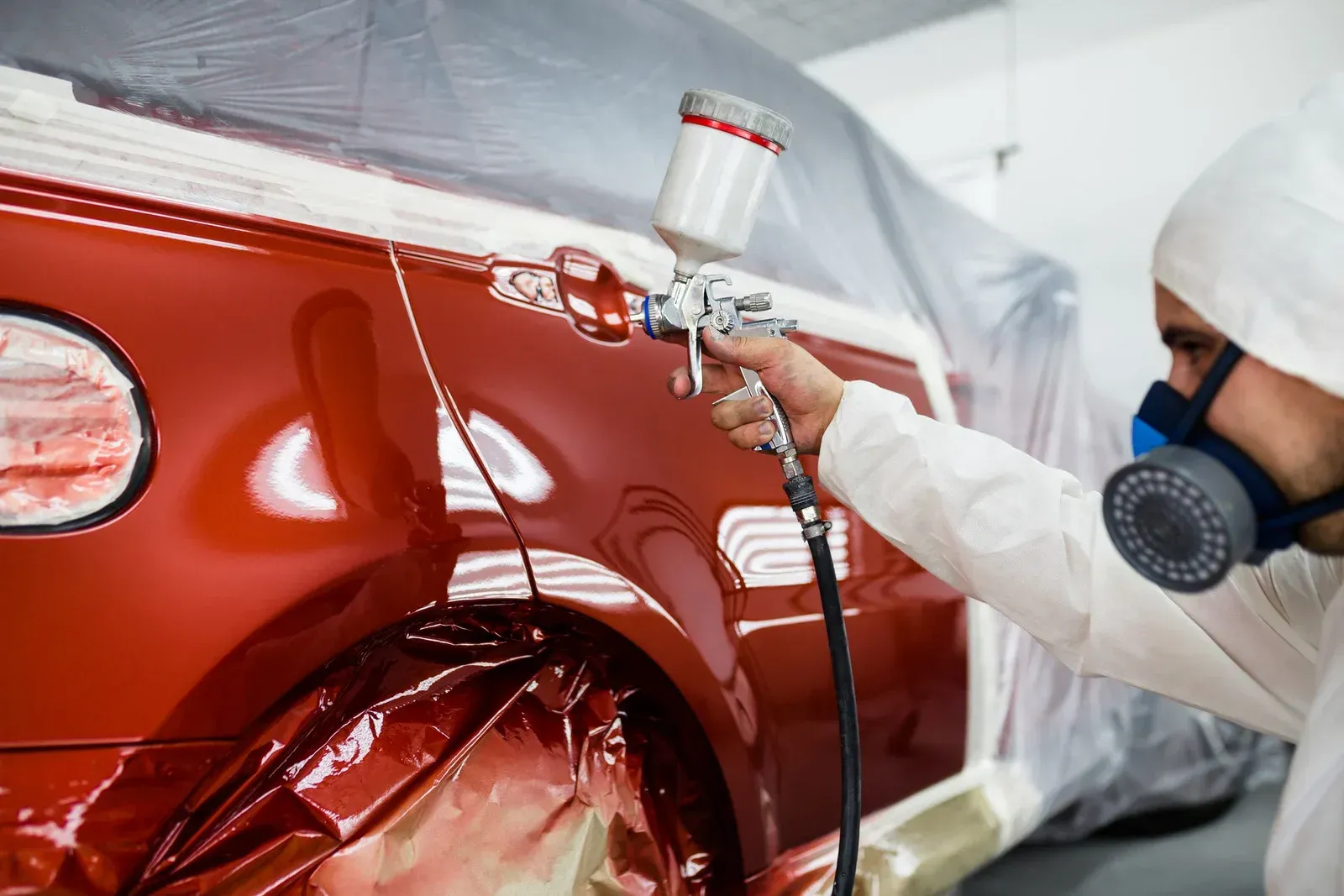 A worker wearing protective gear and blue gloves uses a paint spray gun on a car.
