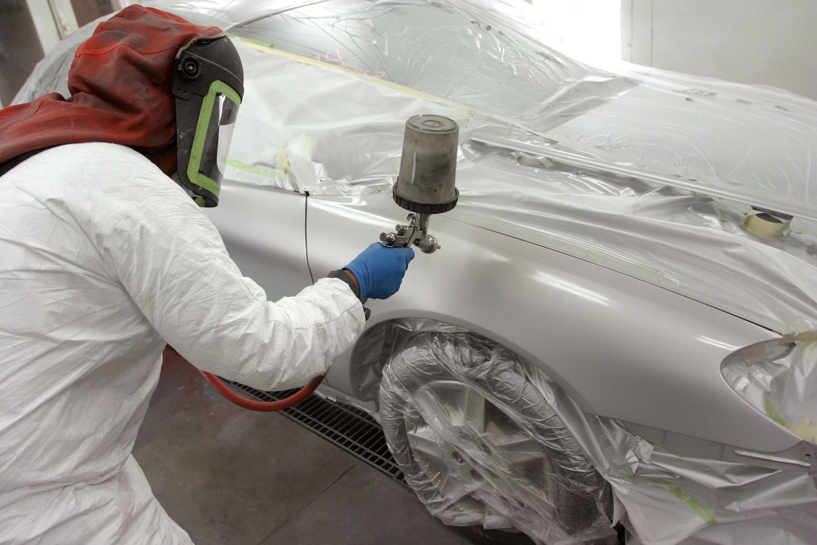 A technician in protective gear uses a spray gun to paint the fender of a car covered in plastic masking film.