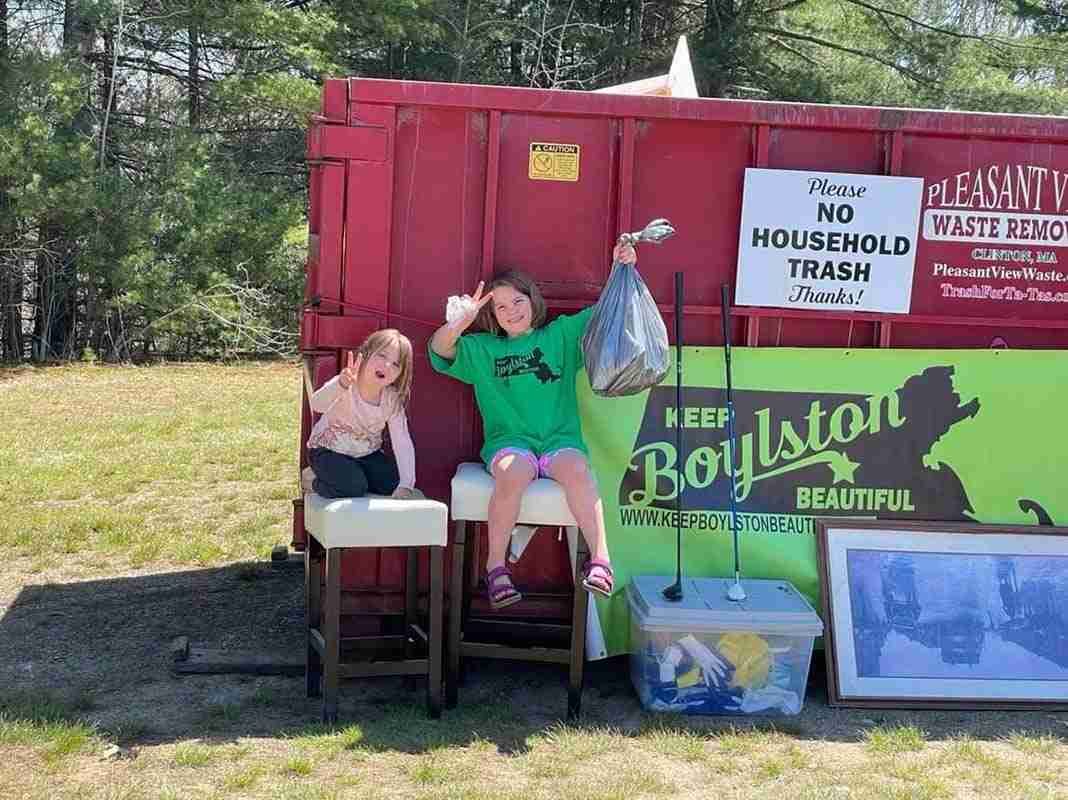 Two young girls are sitting on a bench in front of a dumpster.