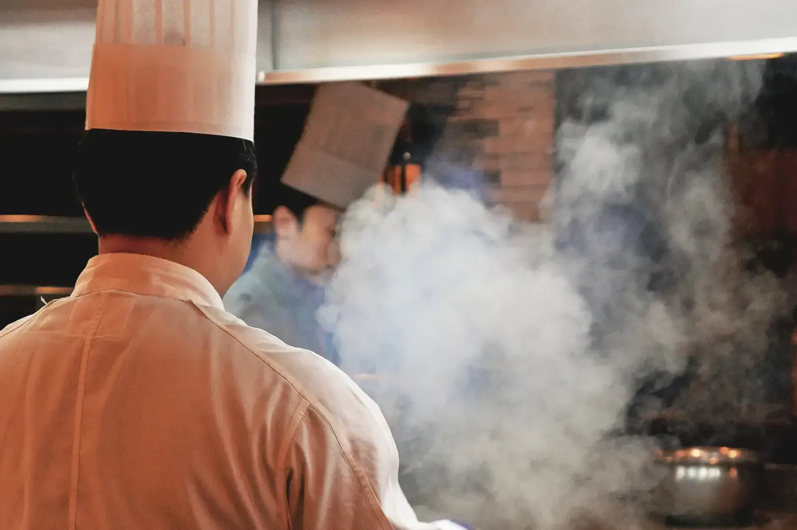 A male chef facing backwards in kitchen area with visible smoke from cooking pan