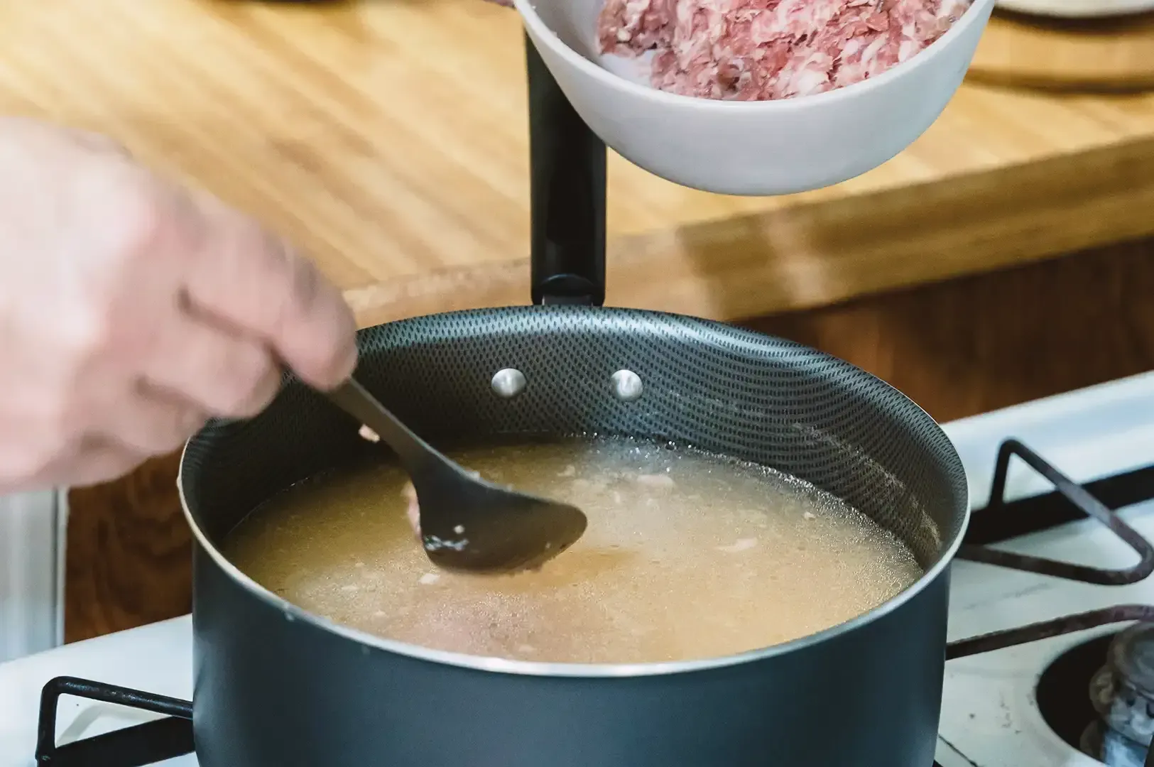 Hand holding a spoon pouring pieces of meat over cooking broth in a black steel pot