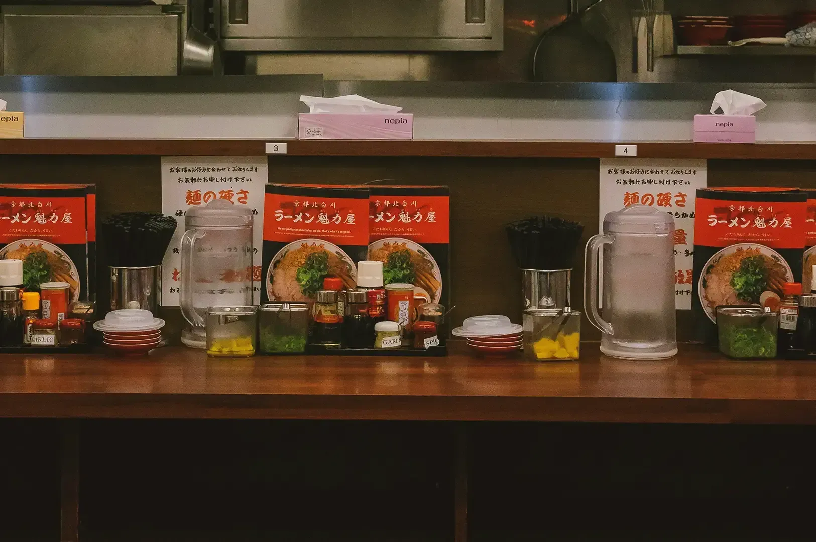 Wooden surface ramen counter filled with ramen menu book, utensils, condiments and water pitcher