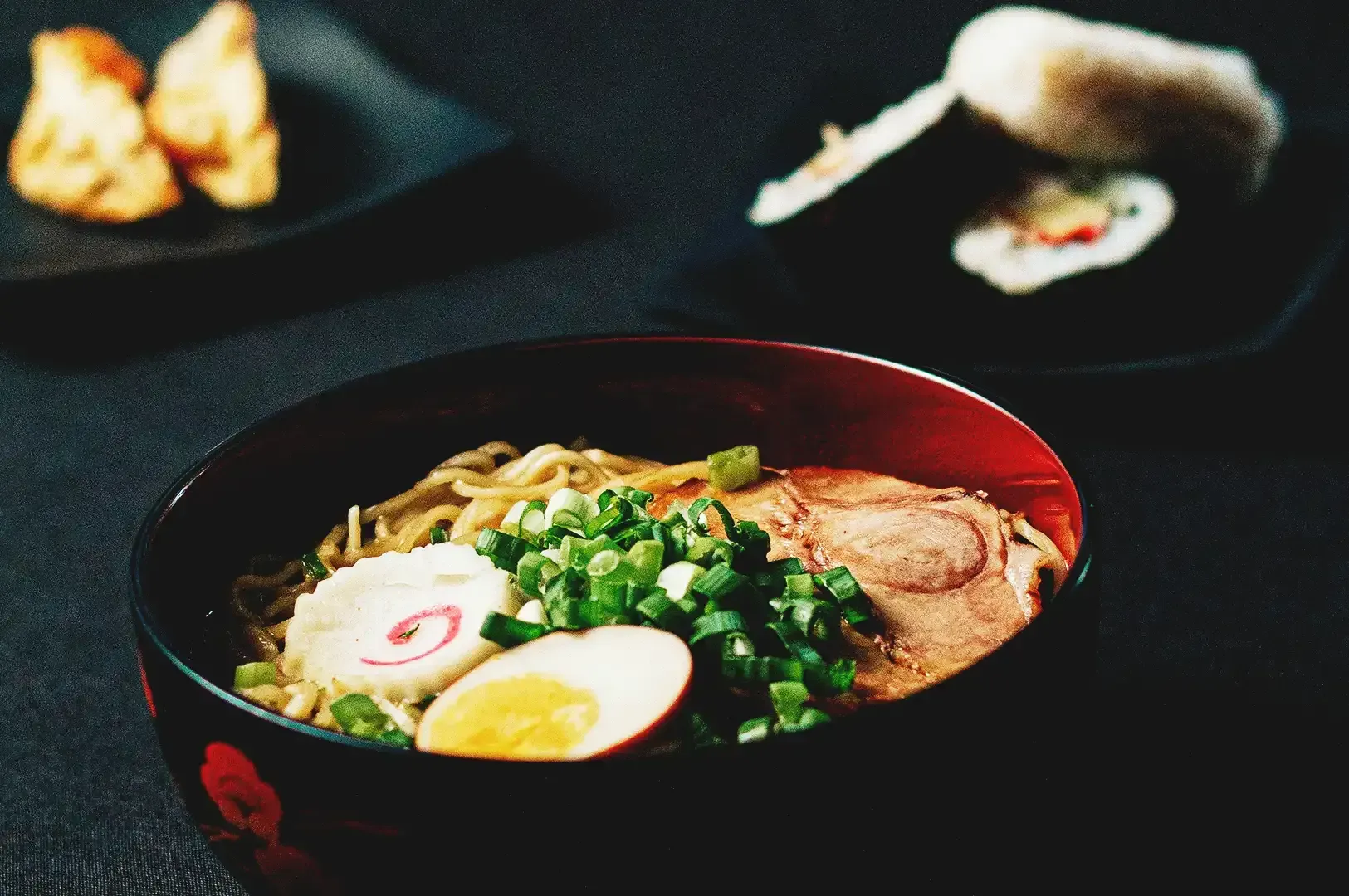 Ramen bowl filled with green onions, noodles, egg and pork meat with blurred sushi and dumpling