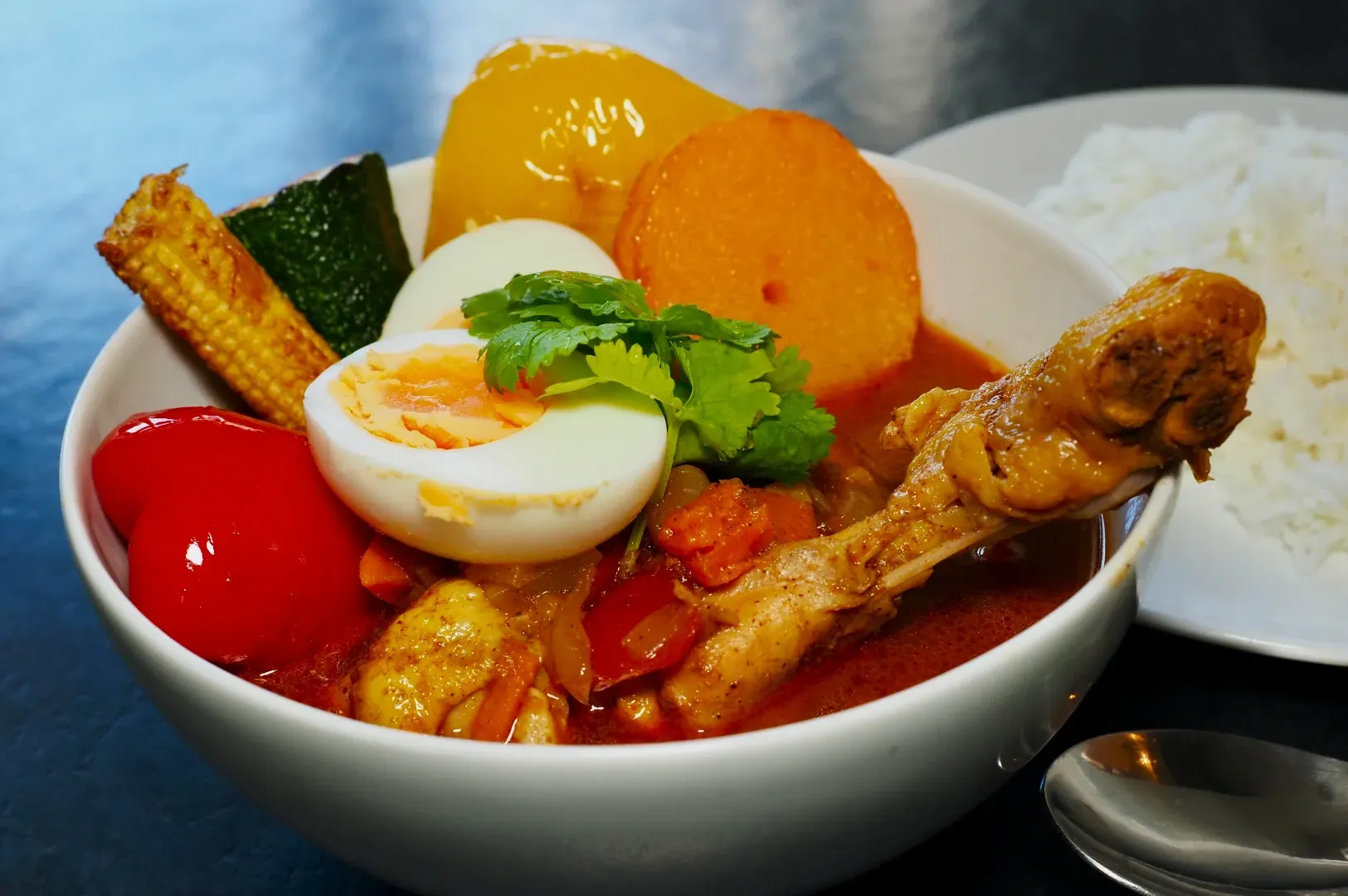 A vibrant bowl of curry featuring a chicken drumstick, boiled egg, assorted colorful vegetables, and topped with cilantro. A side of white rice complements the dish.