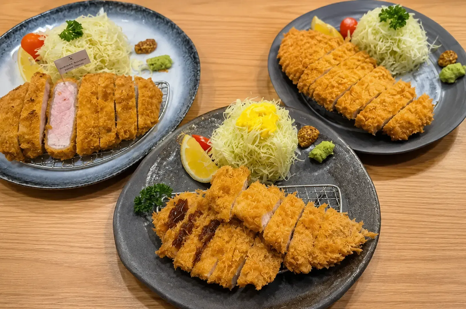 Three plates of crispy tonkatsu pork cutlets served with shredded cabbage, lemon wedges, and garnish