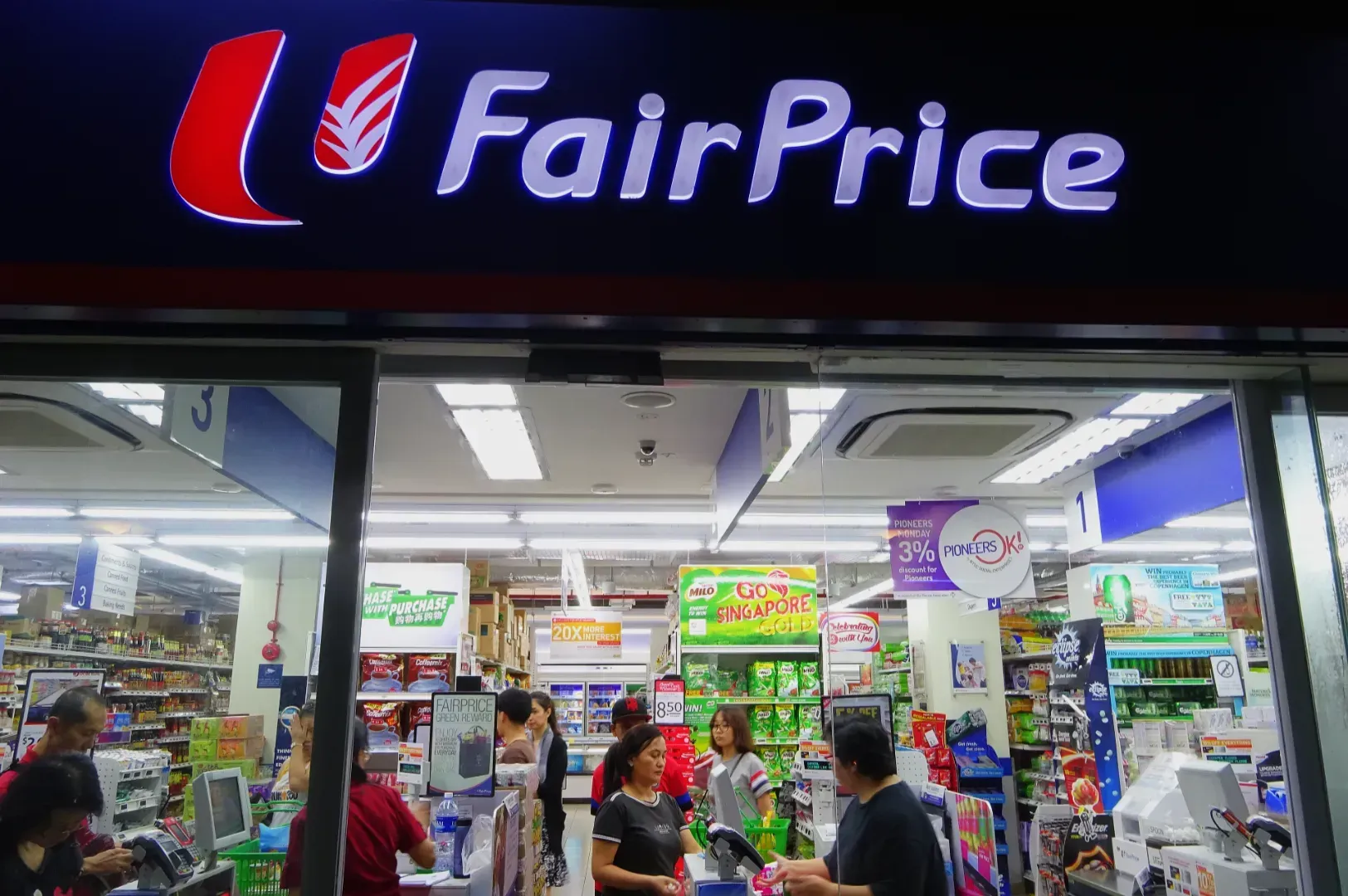 Exterior of a brightly lit FairPrice supermarket at night. Shoppers are seen inside near the checkout counters. The atmosphere is busy and lively.