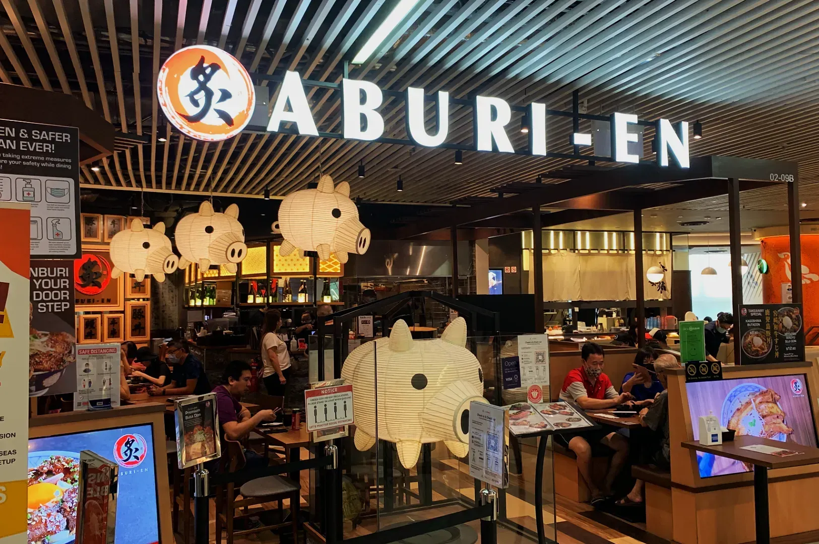 Restaurant entrance of Aburi-En, featuring hanging pig lanterns and patrons dining inside. Warm lighting and cozy ambiance suggest a relaxed dining experience.