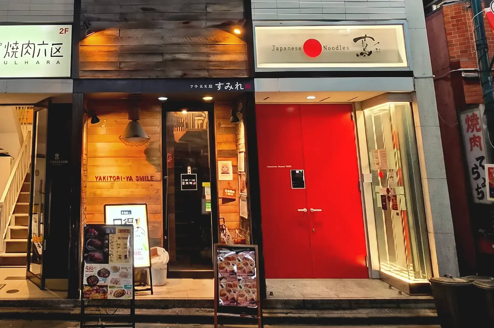 Red door with beige lit signage of Tsuta along with other Japanese food stores