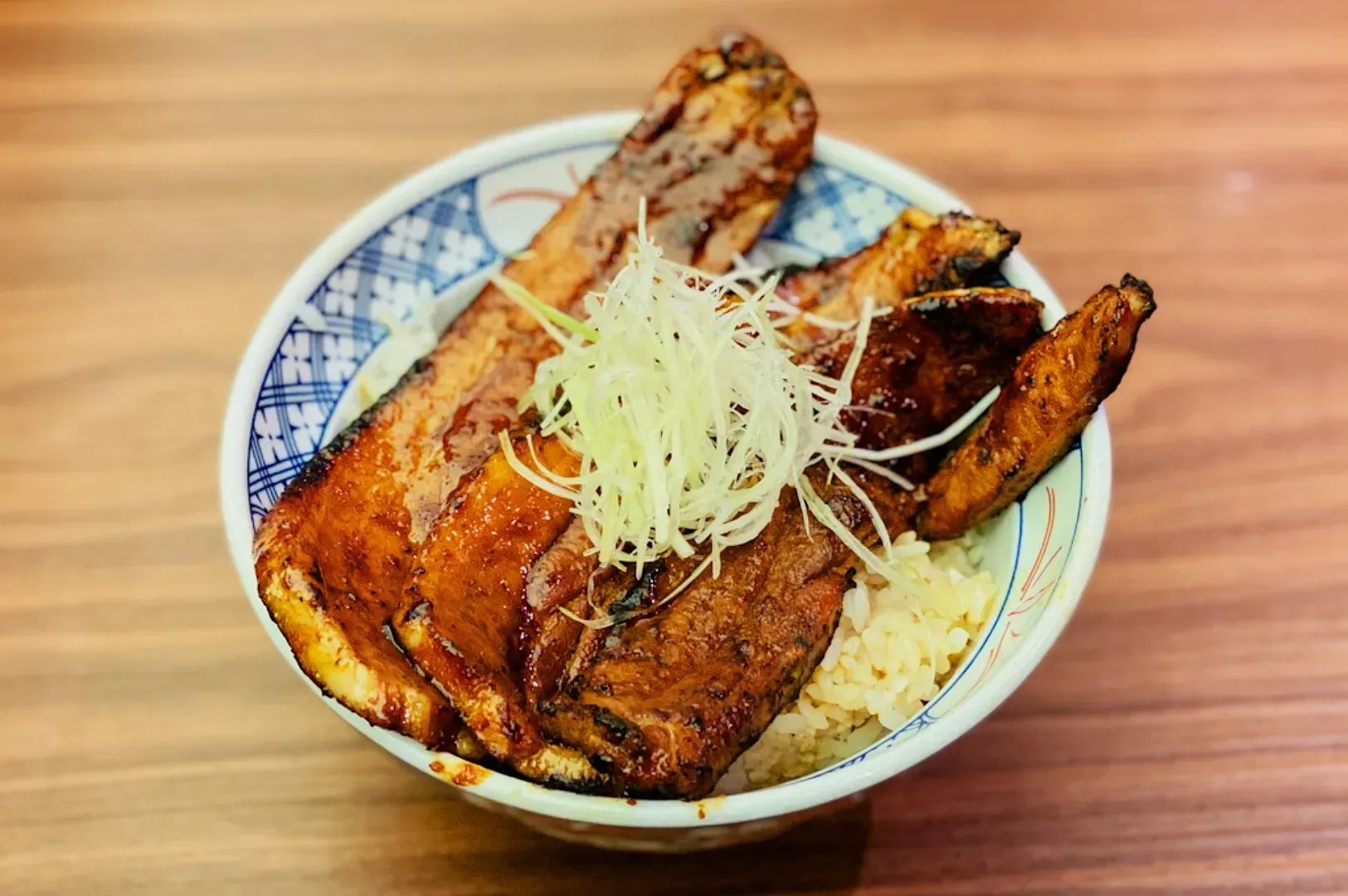 A bowl filled with glazed grilled eel filets over steamed rice, topped with thinly sliced green onions. The setting is a wooden table, conveying a cozy dining atmosphere.