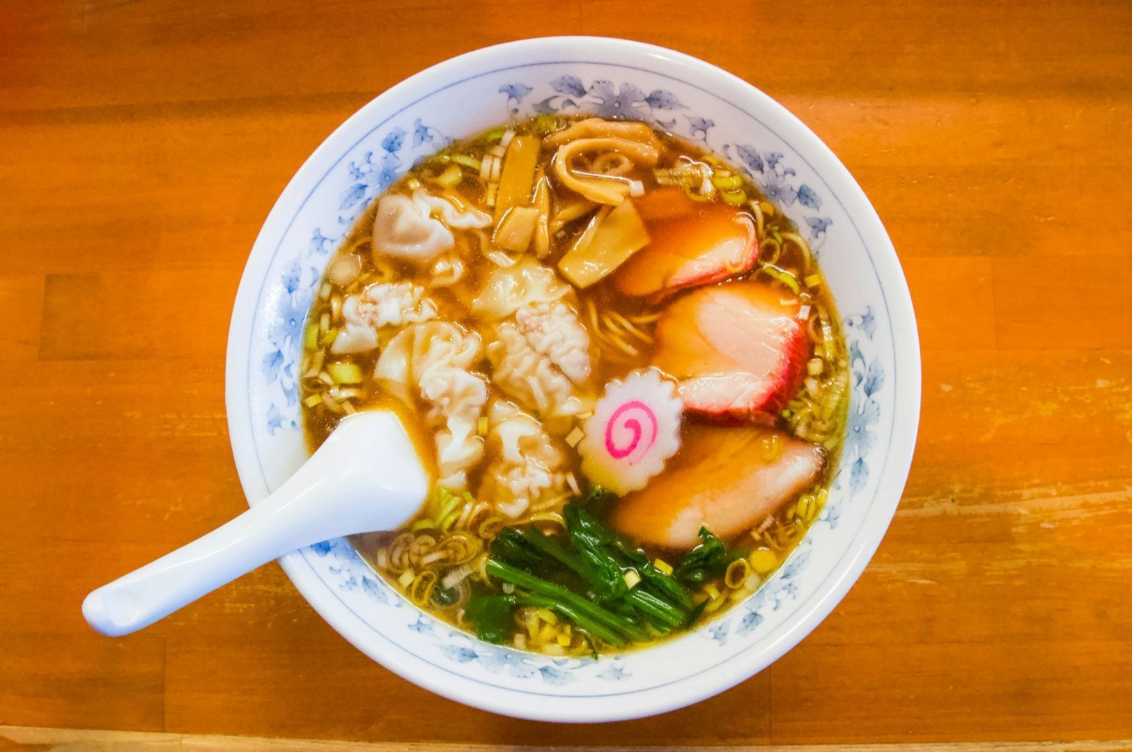 A bowl of ramen on a wooden table, featuring sliced meat, bamboo shoots, a pink spiral fish cake, dumplings, and greens, with a ceramic spoon.