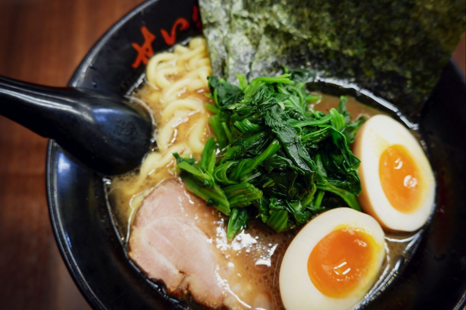 A bowl of ramen with rich broth, topped with sliced pork, spinach, two soft-boiled eggs, and nori in a black bowl, conveying warmth and comfort.
