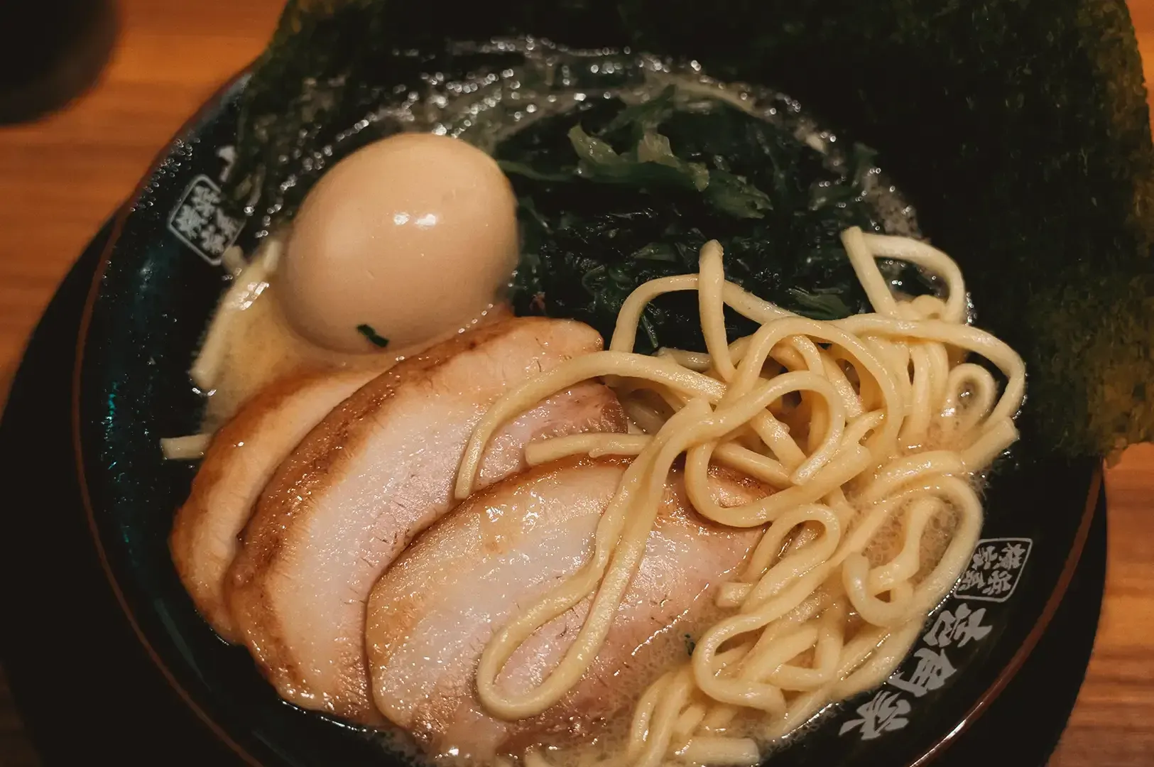 Close up of thick ramen noodles, three pork meat slices, egg, boiled vegetables and seaweed sheets on the side in a black bowl