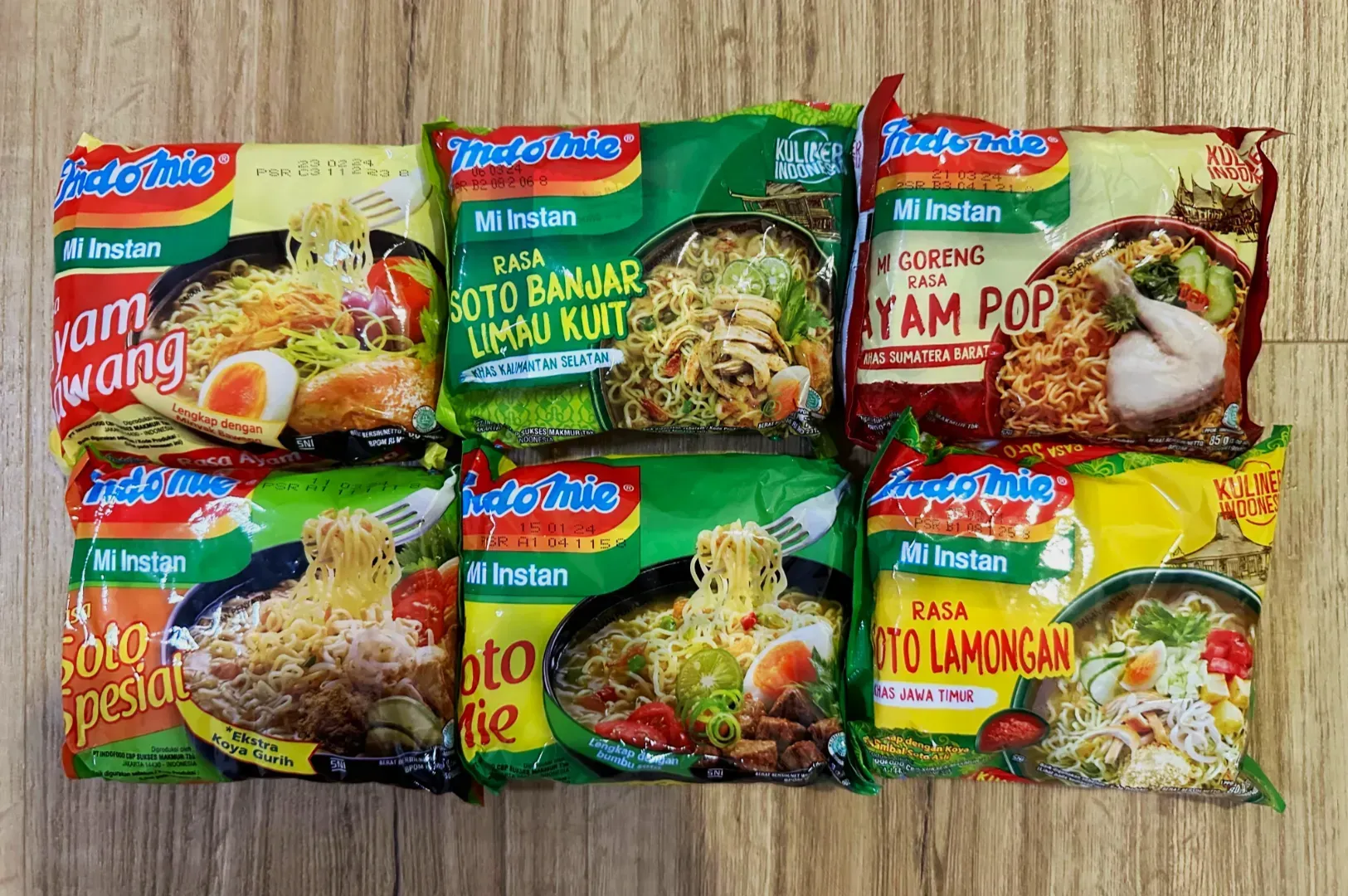 A variety of Indomie instant noodle packages, featuring different flavors, are displayed on a wooden surface. The colorful packaging shows appetizing images of the noodle dishes, depicting an assortment of ingredients like boiled eggs and vegetables.