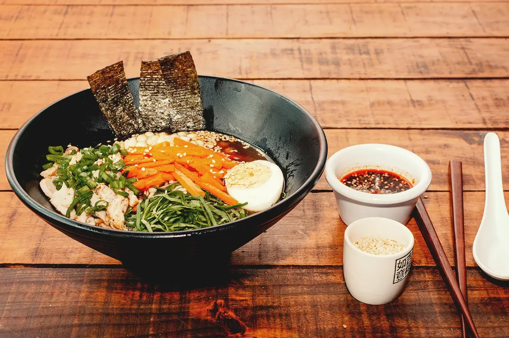 A bowl of ramen topped with veggies, meat and egg and extra seasonings on the side