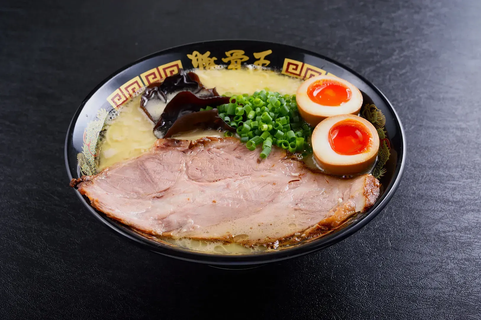 A bowl of ramen with rich broth, topped with sliced pork, a halved soft-boiled egg, green onions, and mushrooms, on a dark surface.