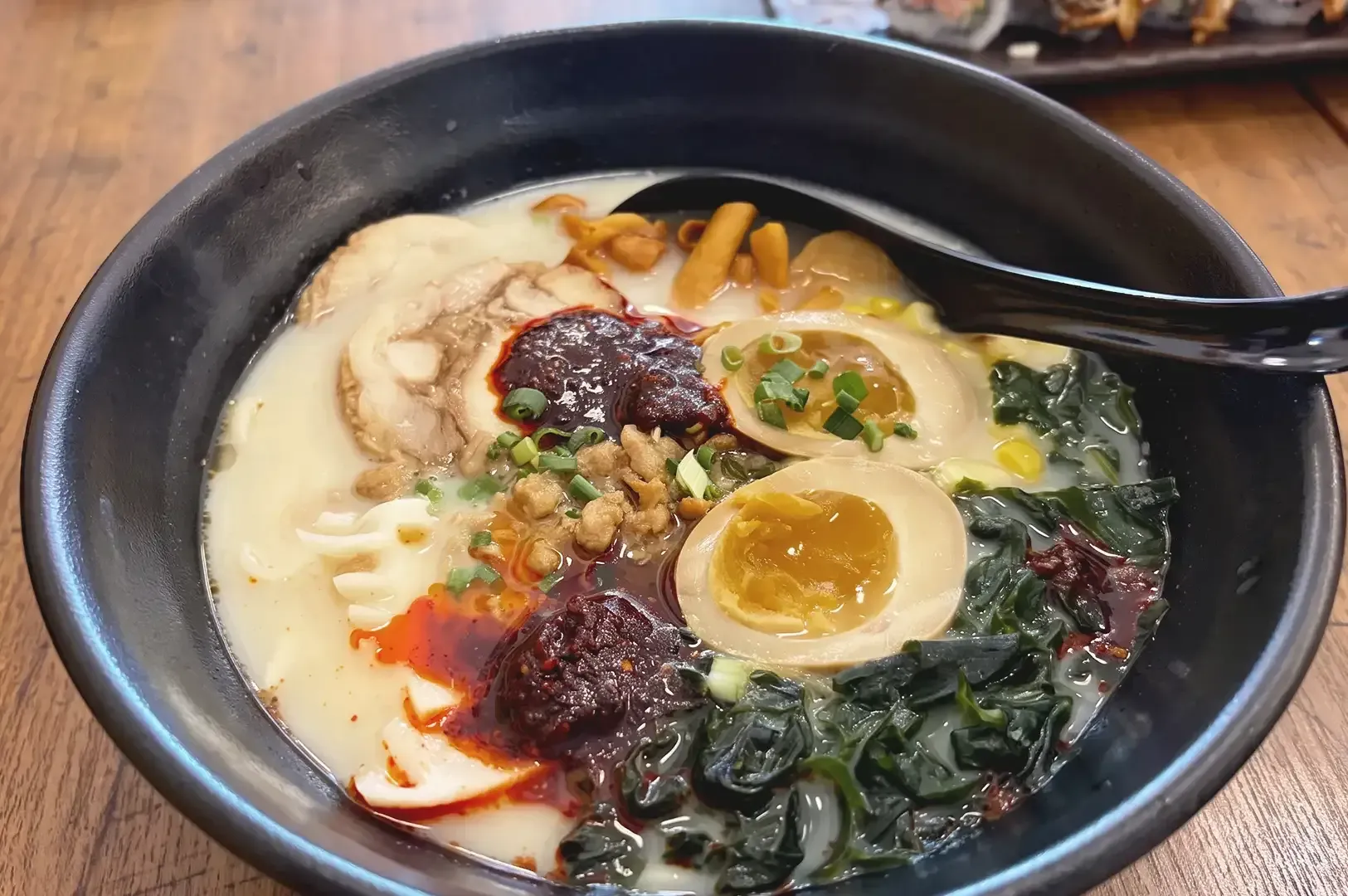 Creamy ramen broth topped with eggs, chili sauce, vegetables and pork meat slightly scooped by a black spoon