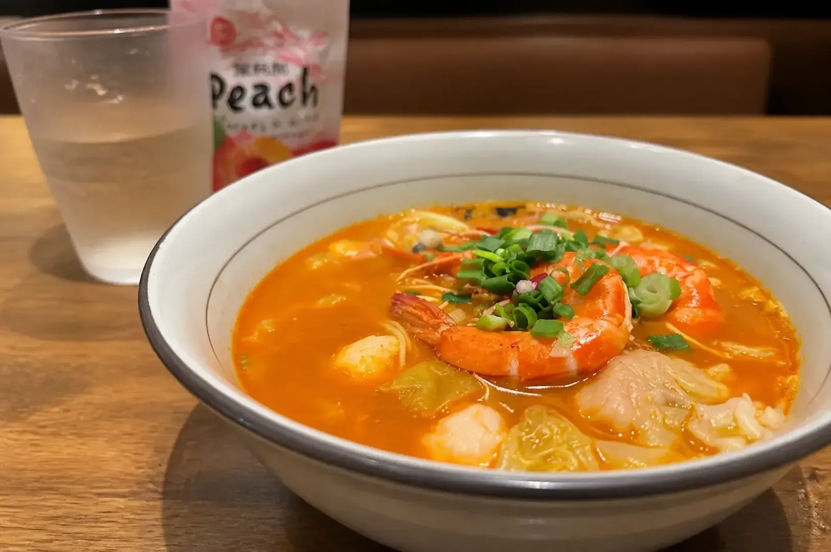 Bowl of shrimp ramen topped with chopped green onions on a wooden table, accompanied by a peach drink pouch
