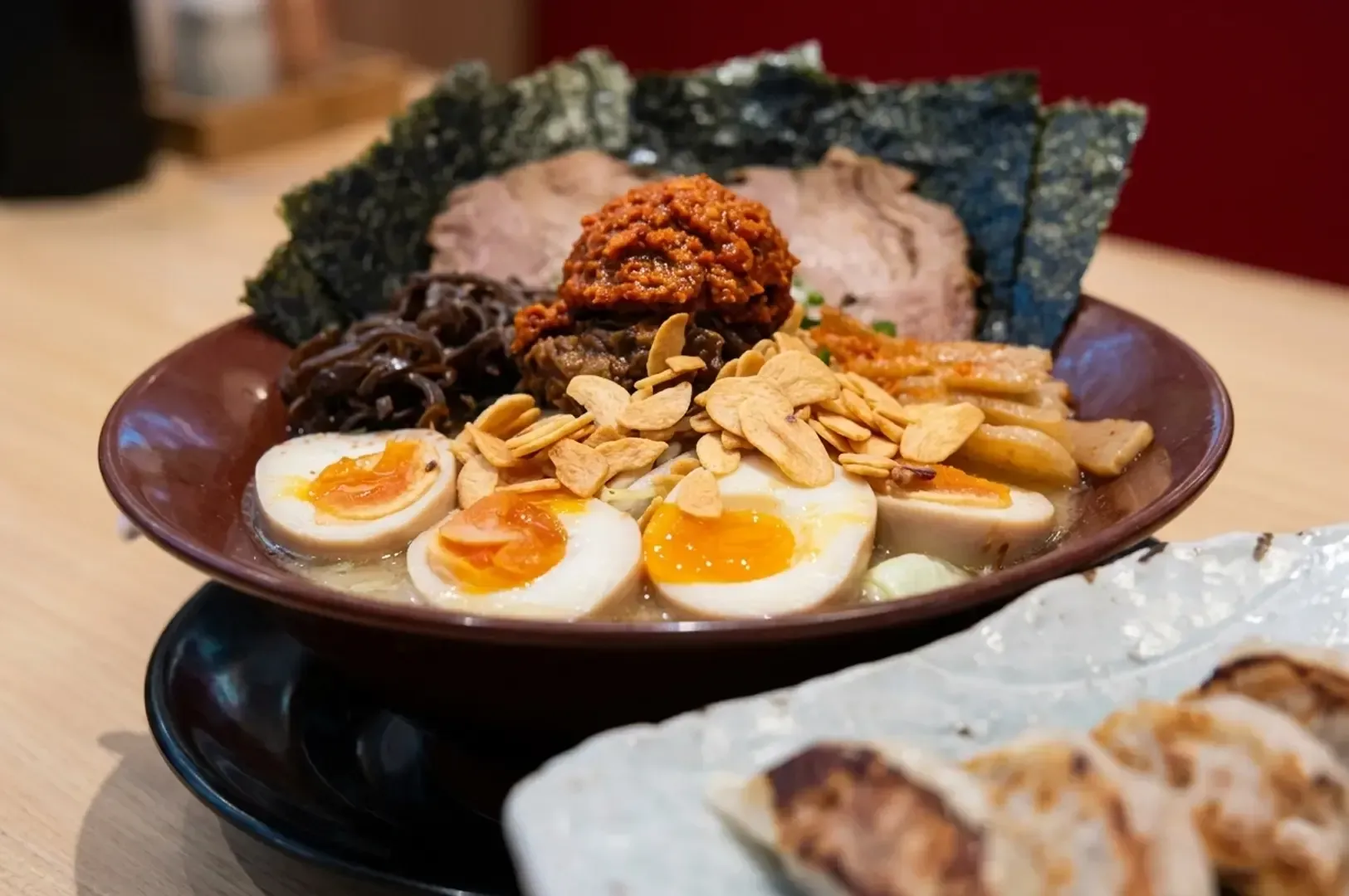 Loaded ramen bowl with multiple eggs, pork, garlic chips, and spicy miso topping next to gyoza.
