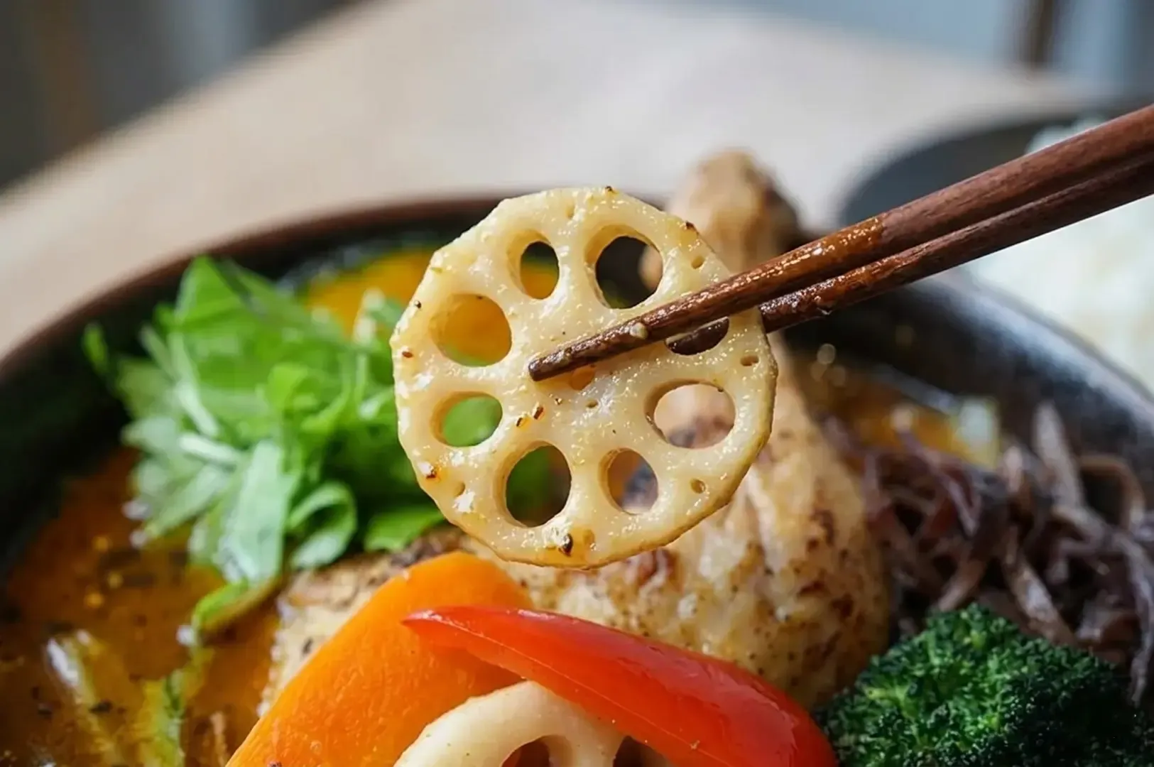 Close-up of wooden chopsticks holding a slice of lotus root over a bowl of Japanese soup curry with chicken and vegetables blurred in the background