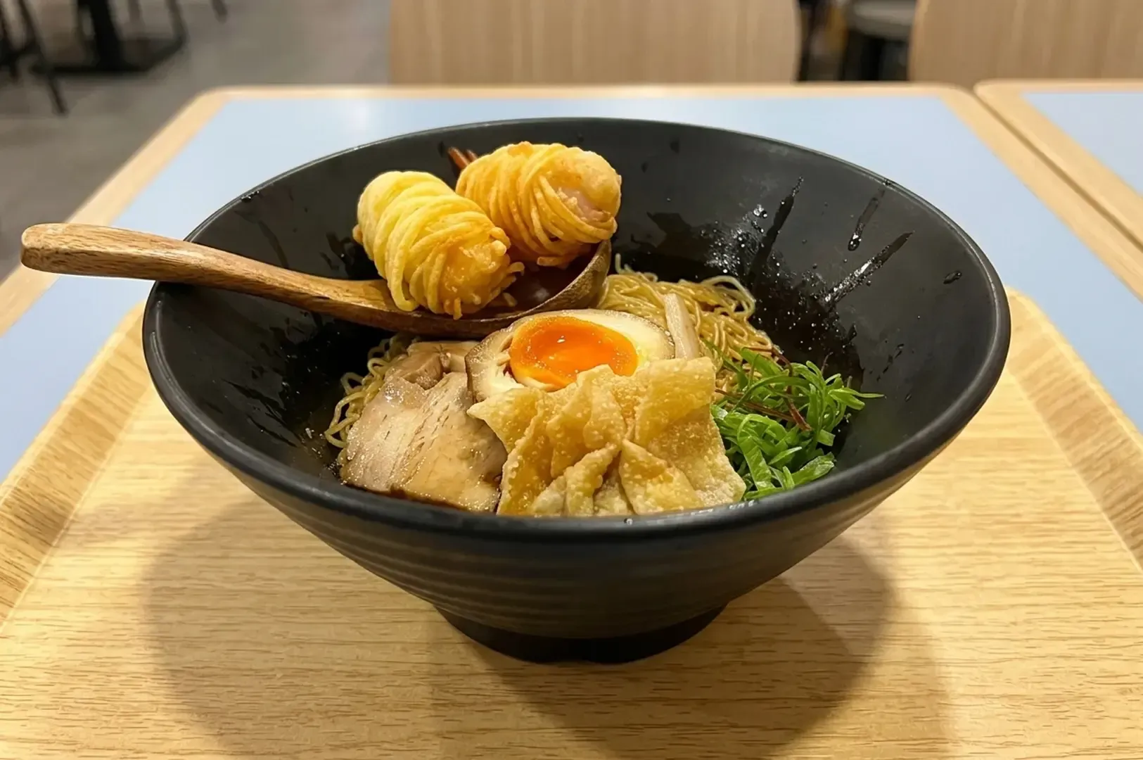 Black bowl of ramen with pork, egg, wontons, and spiral fried prawns on a wooden spoon.