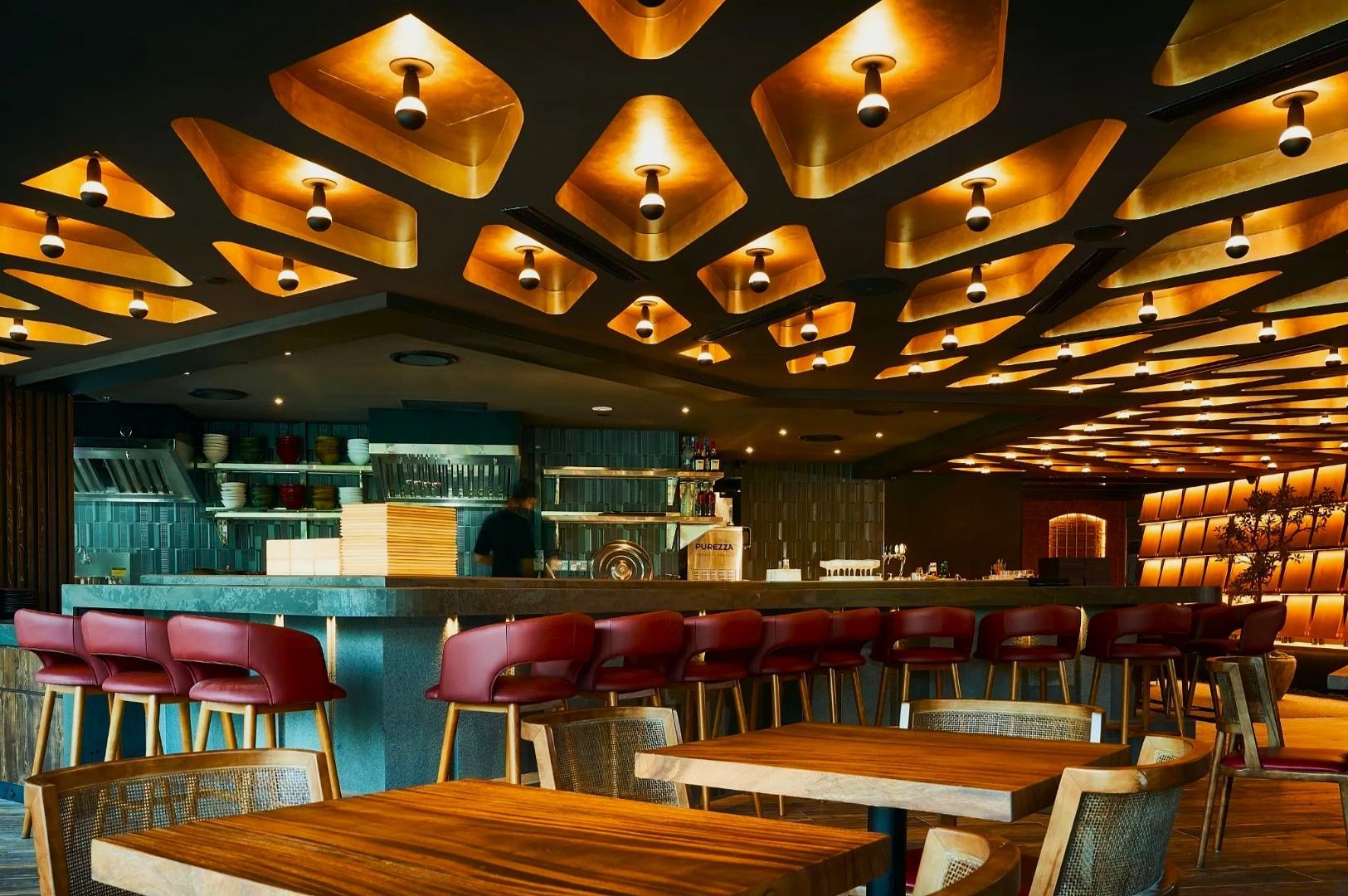 Stylish restaurant interior with geometric ceiling lights, a sleek bar with red stools, and wooden tables and chairs. Warm, inviting atmosphere.