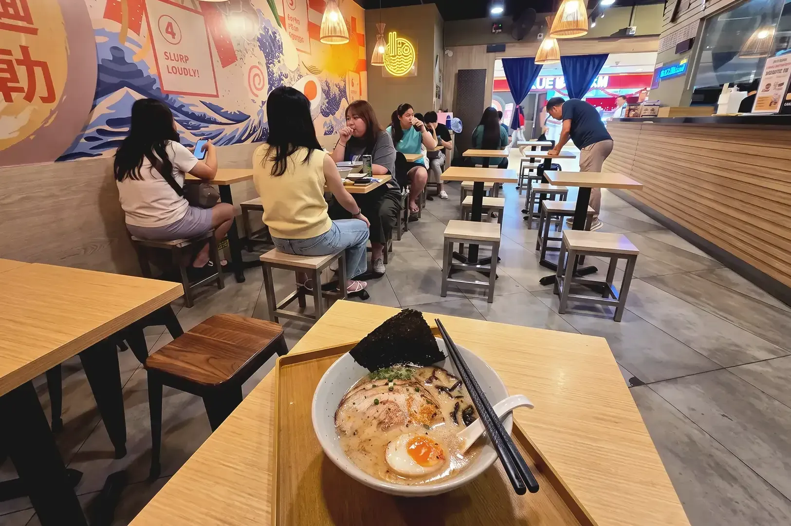 Orange, blue and yellow motif Japanese decorated interior of Suparakki Ramen and a bowl of ramen on top of table with people eating around