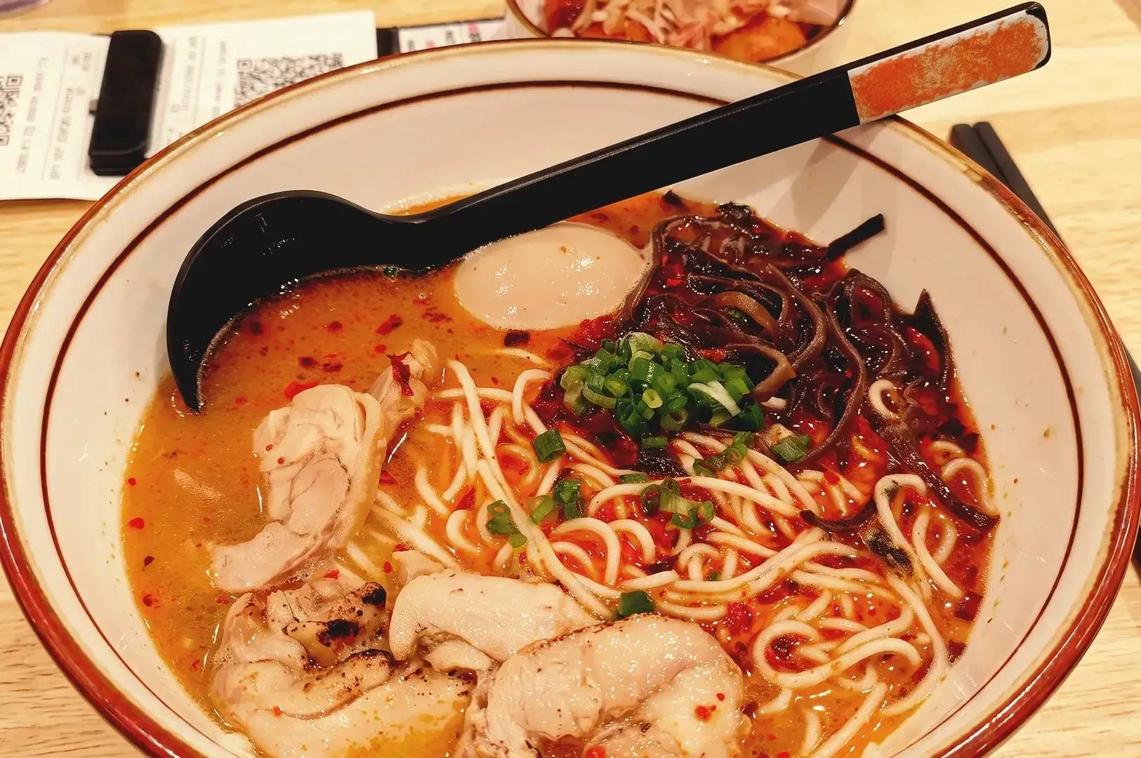 Black spoon slightly scooped on ramen noodles drenched in red ramen broth topped with spring onions, pork meat and black fungus