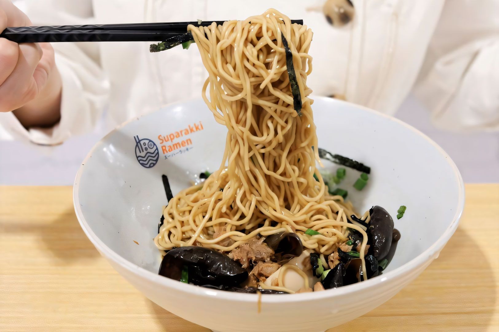 Close-up of black chopsticks lifting a large portion of ramen noodles from a white bowl bearing the Suparakki Ramen logo, showing wood ear mushrooms and toppings below.