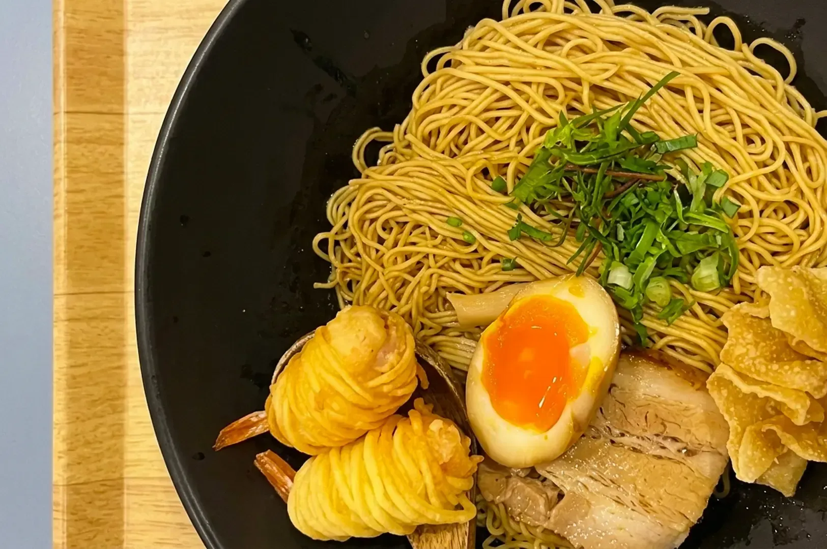 Top-down view of dry noodles with pork, egg, green onions, and fried prawns