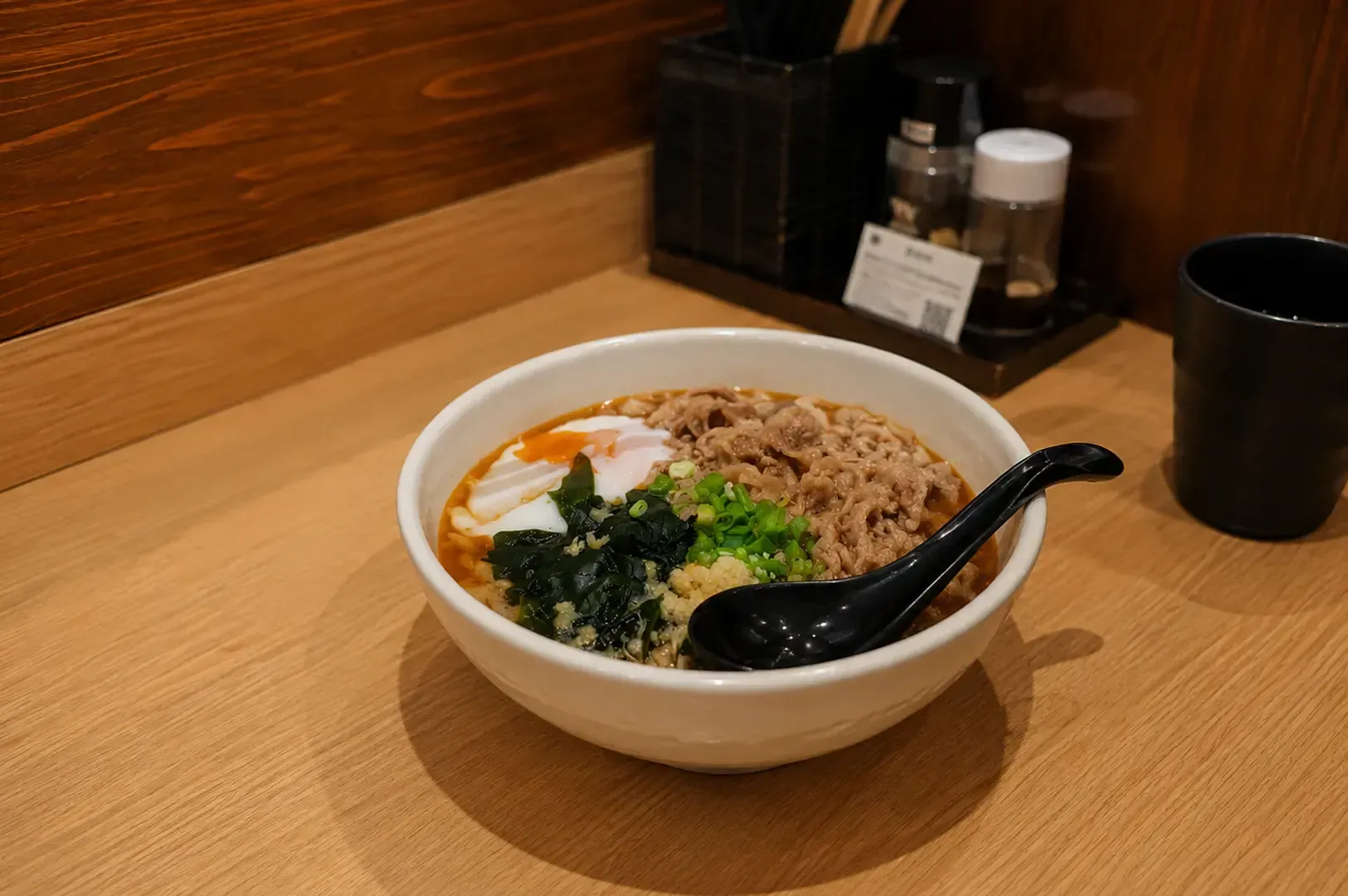 Top-down view of a bowl of udon noodles topped with sliced beef, green onions, minced garlic, and seaweed