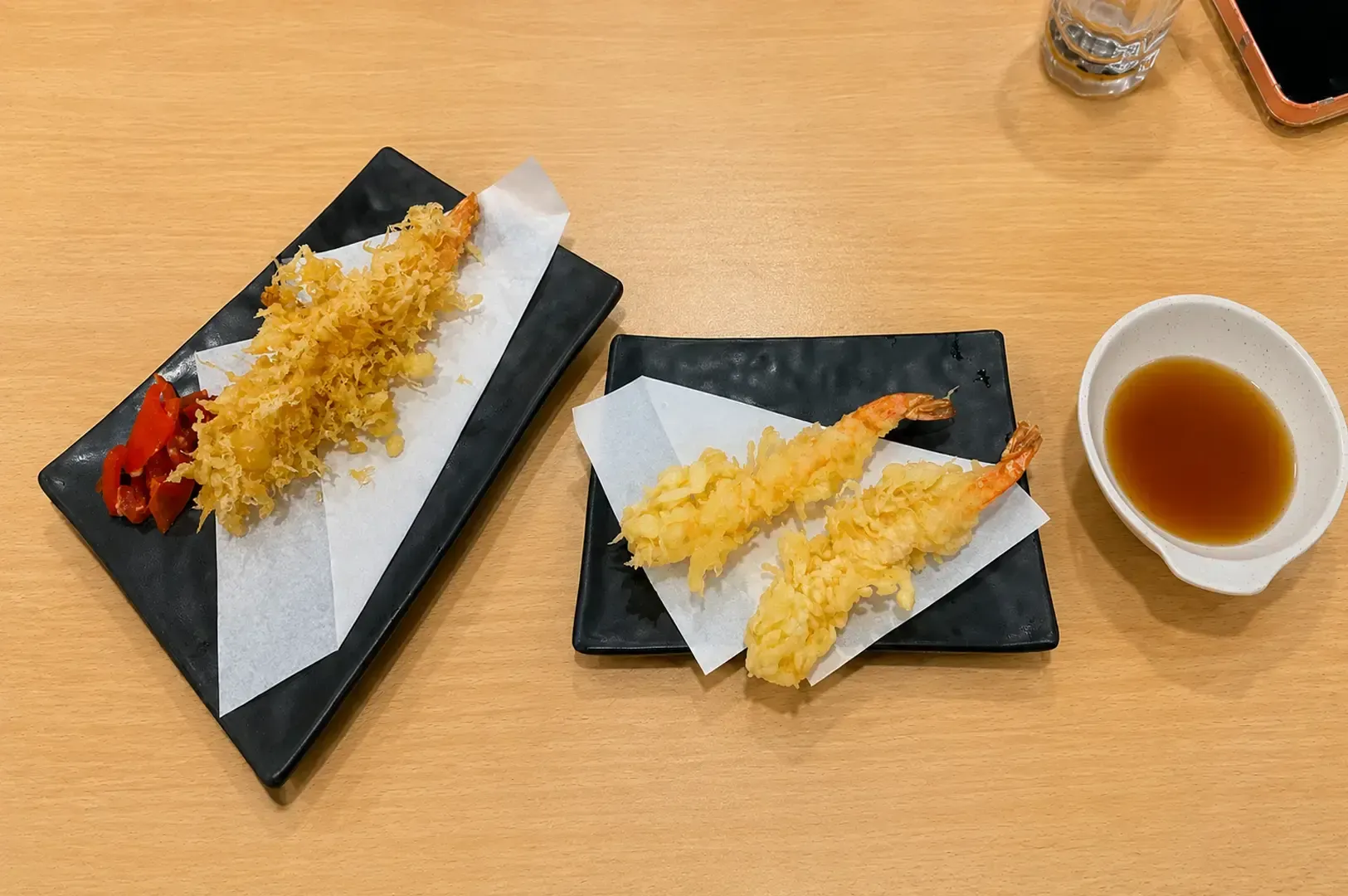 Two black rectangular plates serving golden-brown shrimp tempura and vegetable tempura, accompanied by a small bowl of dipping sauce