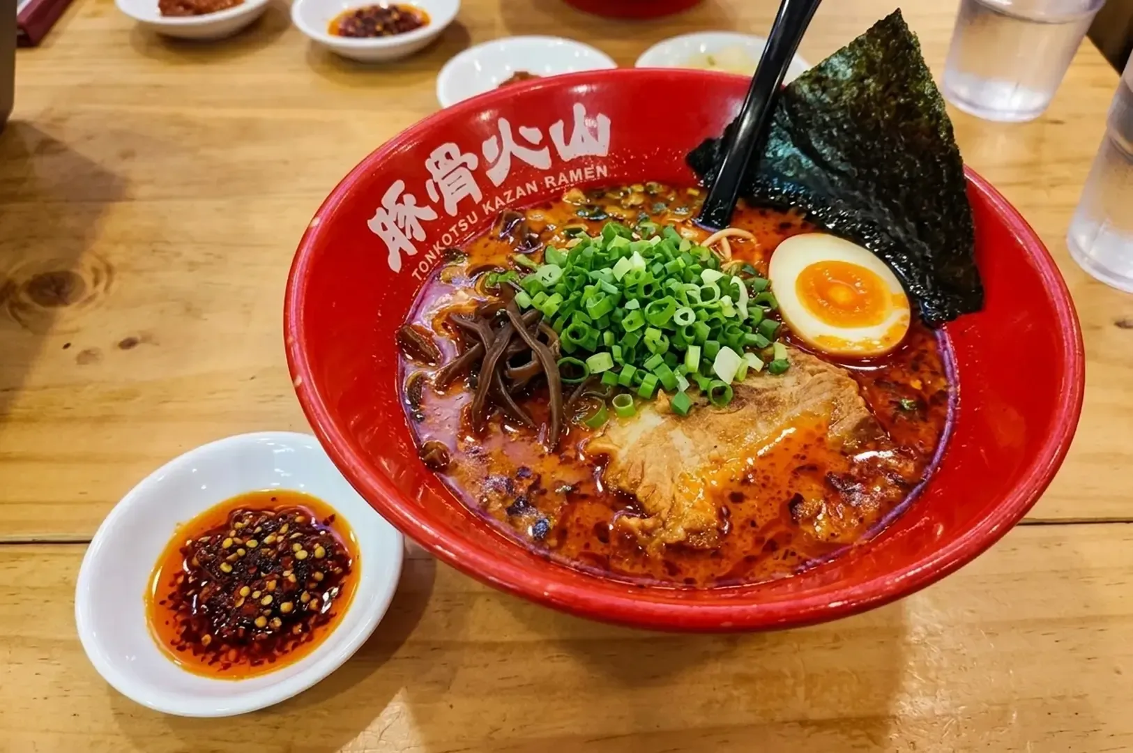 Red bowl of spicy Tonkotsu Kazan Ramen with pork, egg, and chili oil on a wooden table
