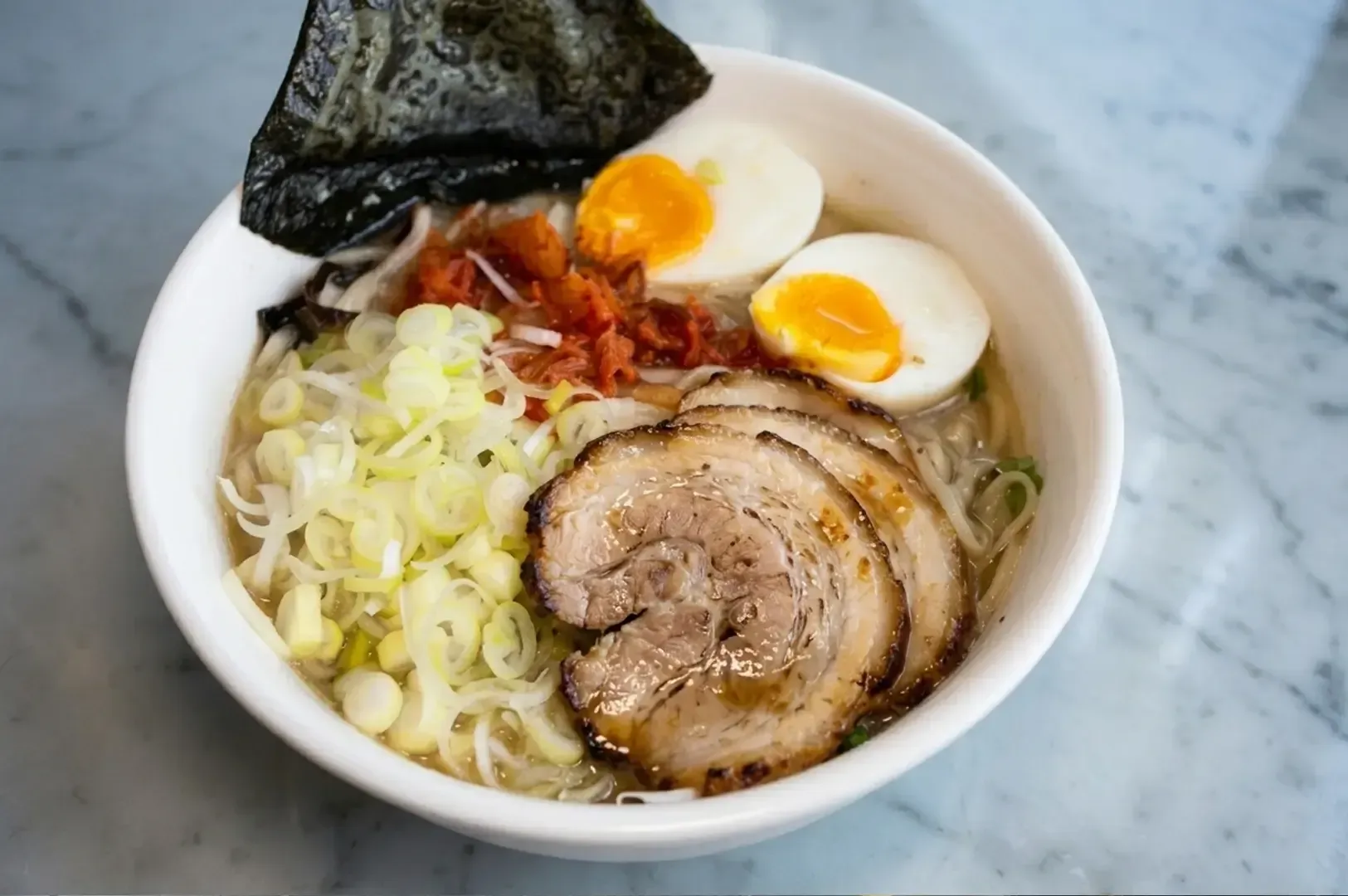 White bowl of ramen topped with chashu pork slices, soft-boiled eggs, nori sheet, and chopped scallions