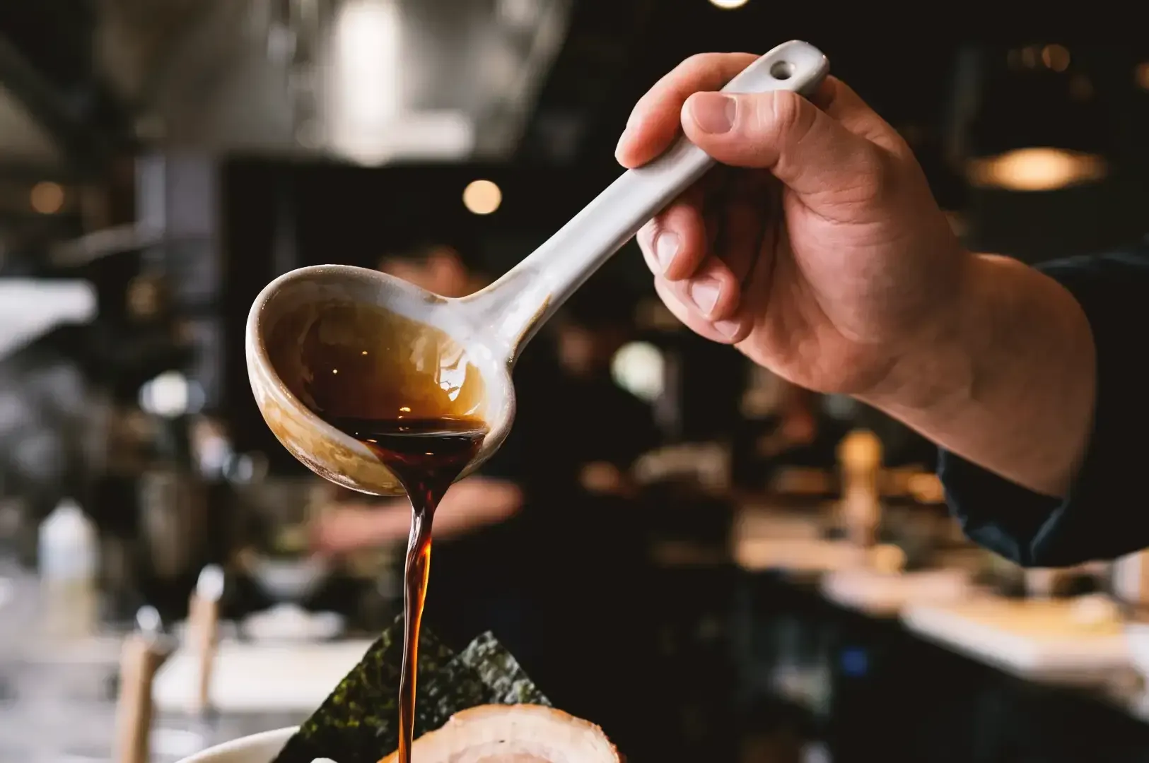 Focused view of hand holding white ladle pouring tare sauce over ramen