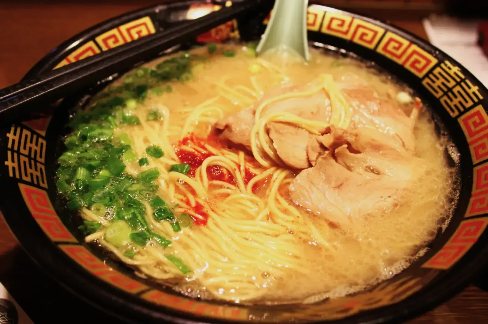 Bowl of ramen with rich broth, topped with sliced pork, green onions, and spicy red sauce. Chopsticks and a spoon rest beside it, creating a comforting vibe.