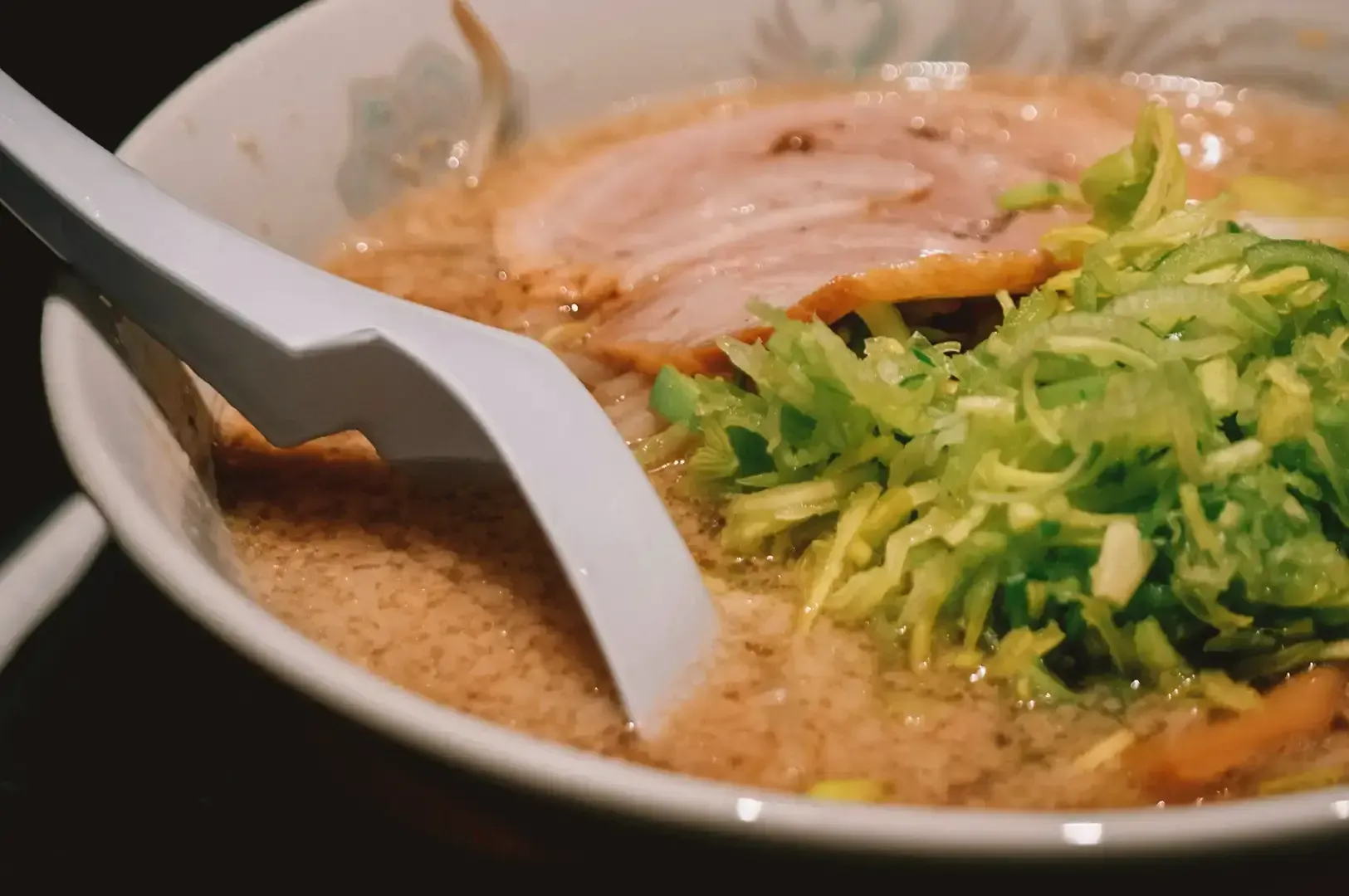 Close up of white spoon dipped in creamy ramen broth with chashu and shredded cabbage as garnish