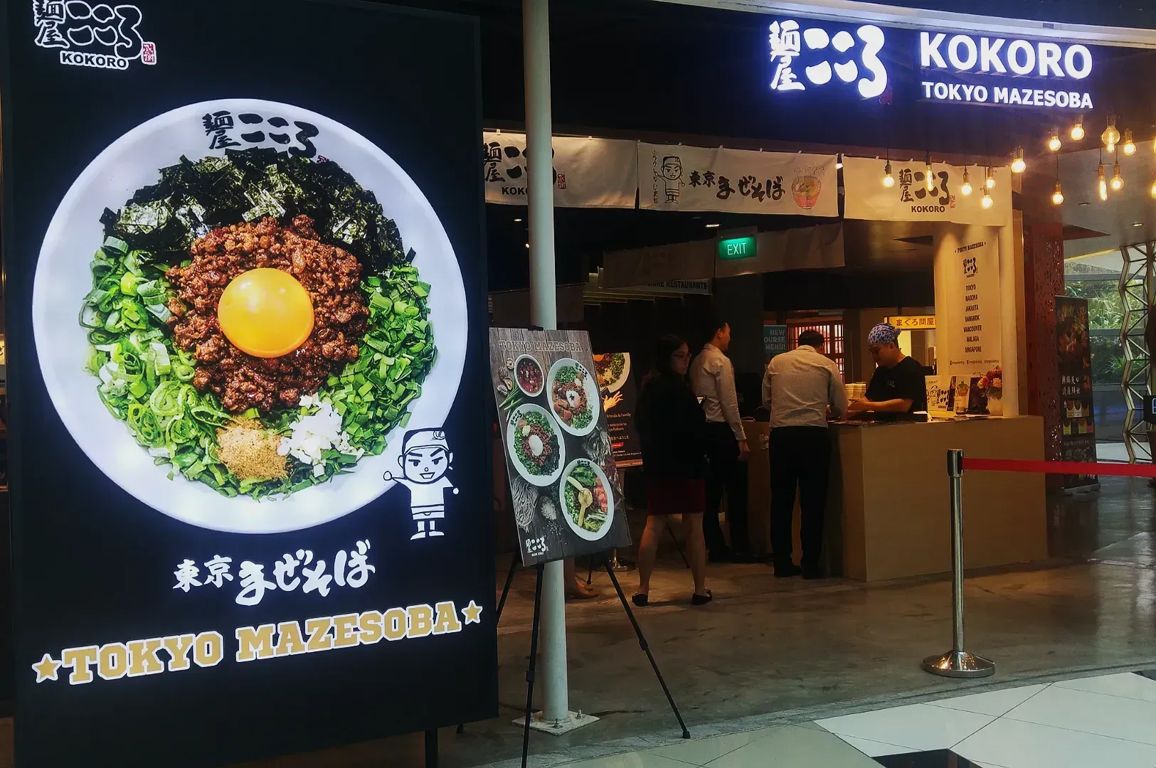 Restaurant entrance with a sign displaying a bowl of mazesoba topped with an egg yolk, greens, and noodles. People are ordering inside Kokoro Tokyo Mazesoba.