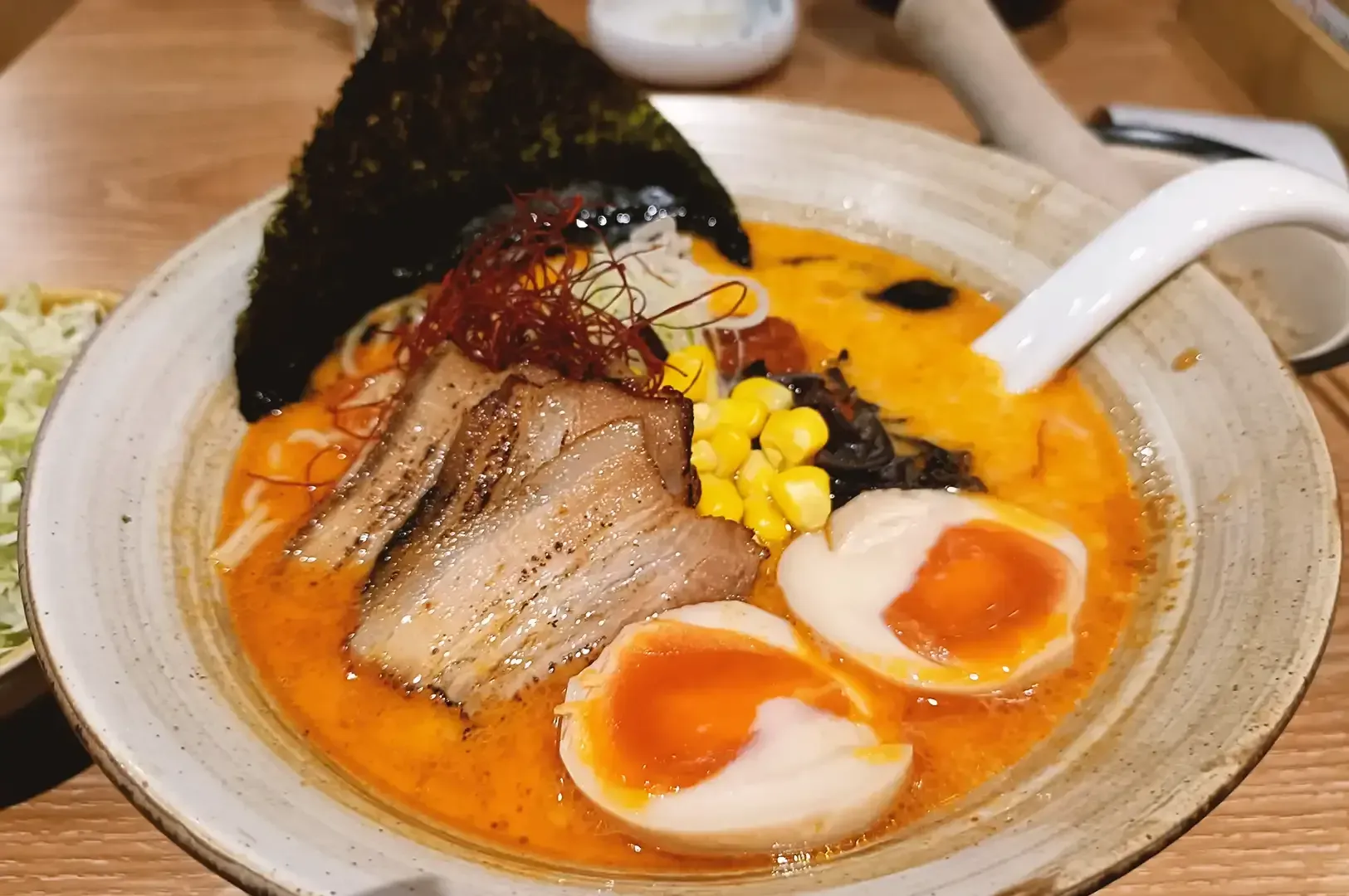 Ramen bowl containing slices of roasted pork, corn, eggs in half, seaweed and ito togarashi