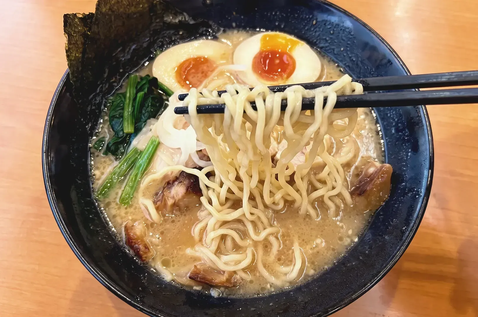 Chopsticks lifting up curly ramen noodles from the broth with eggs, veggues and pork meat