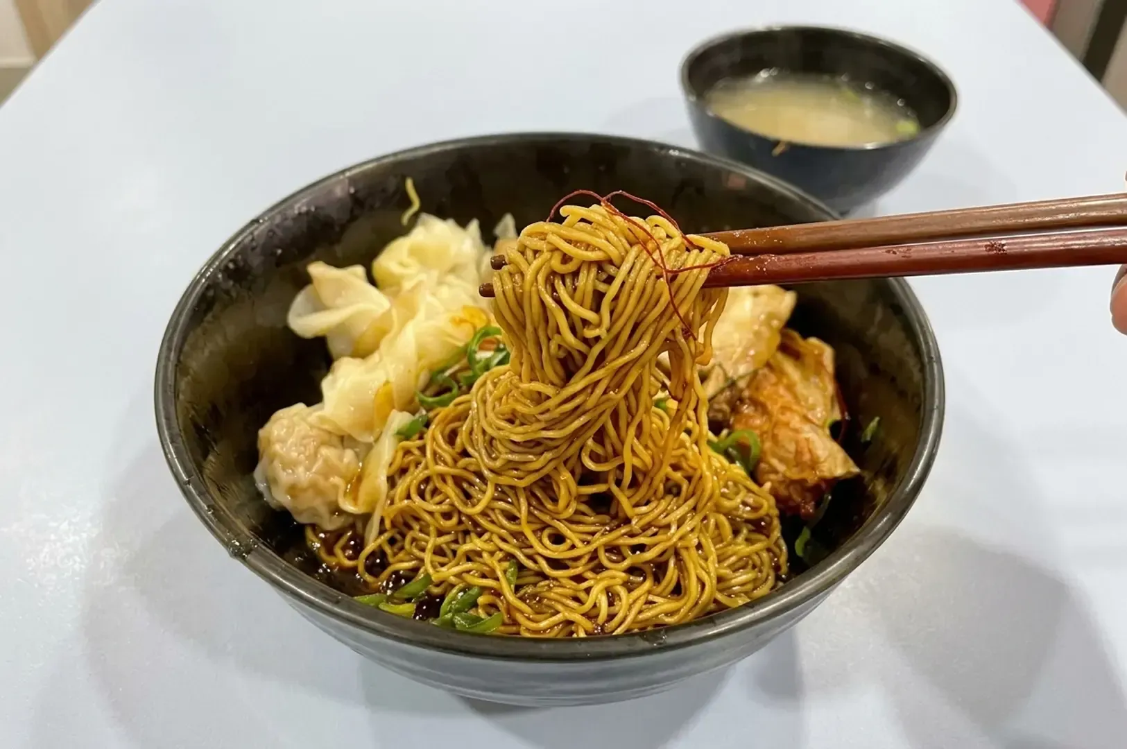 Close-up of chopsticks lifting yellow noodles from a bowl filled with wontons and pork, with soup on the side.
