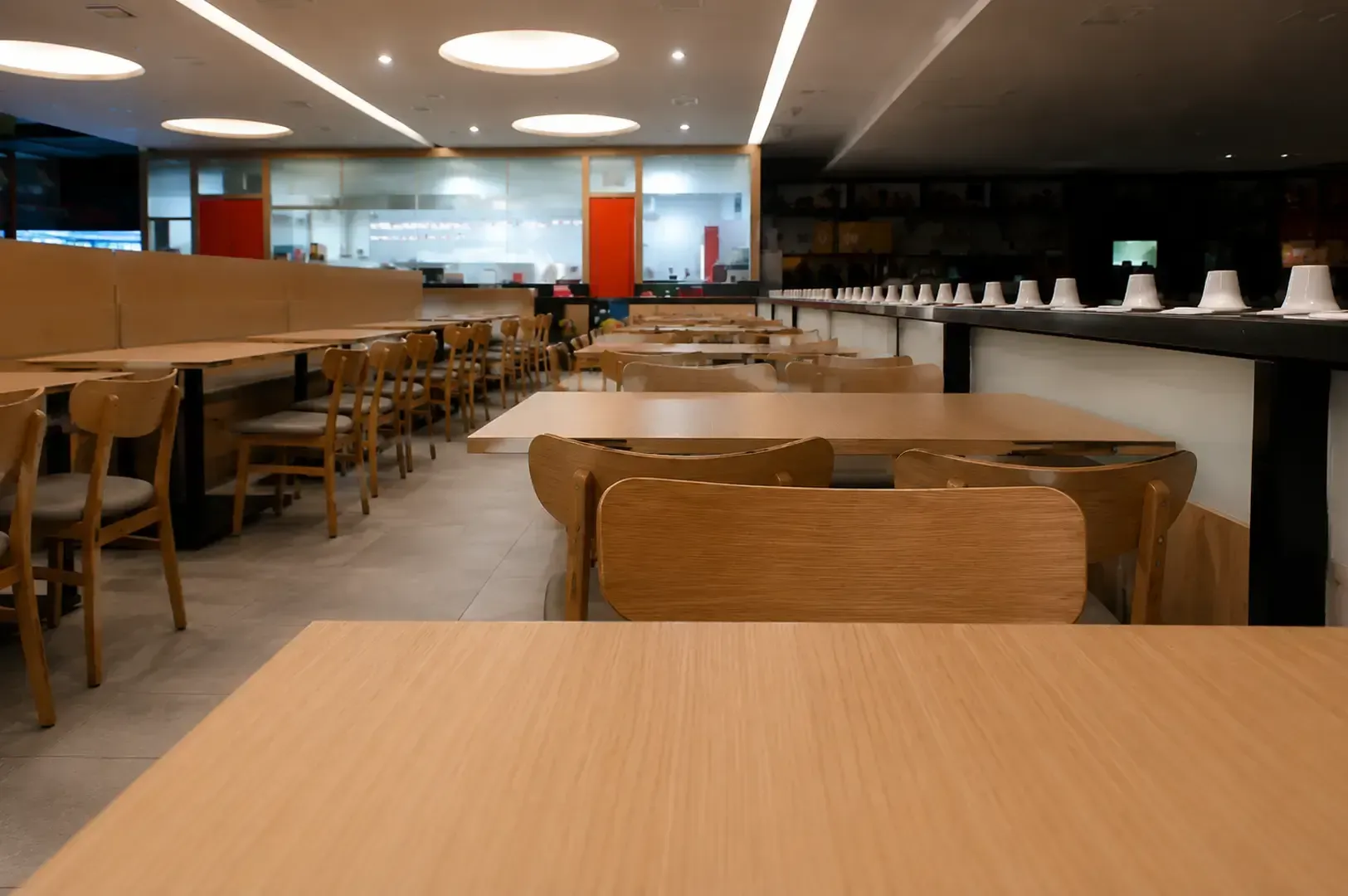 Spacious and modern interior of Tamoya Udon featuring light wooden tables and chairs, with a view of the open kitchen through glass panels in the background