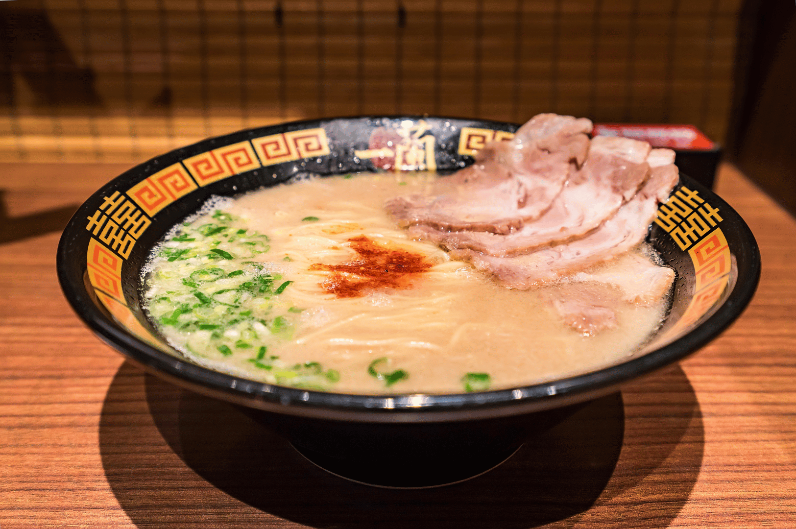 A luxurious bowl of ramen served in a black bowl with red and gold accents, featuring creamy broth, thin noodles, green onions, a central dollop of red seasoning, and neatly arranged slices of pork belly. Set against a wooden table with a bamboo screen backdrop, the scene evokes traditional Japanese dining aesthetics, spotlighting the elegance and depth of flavor found in Singapore’s premium ramen experiences.